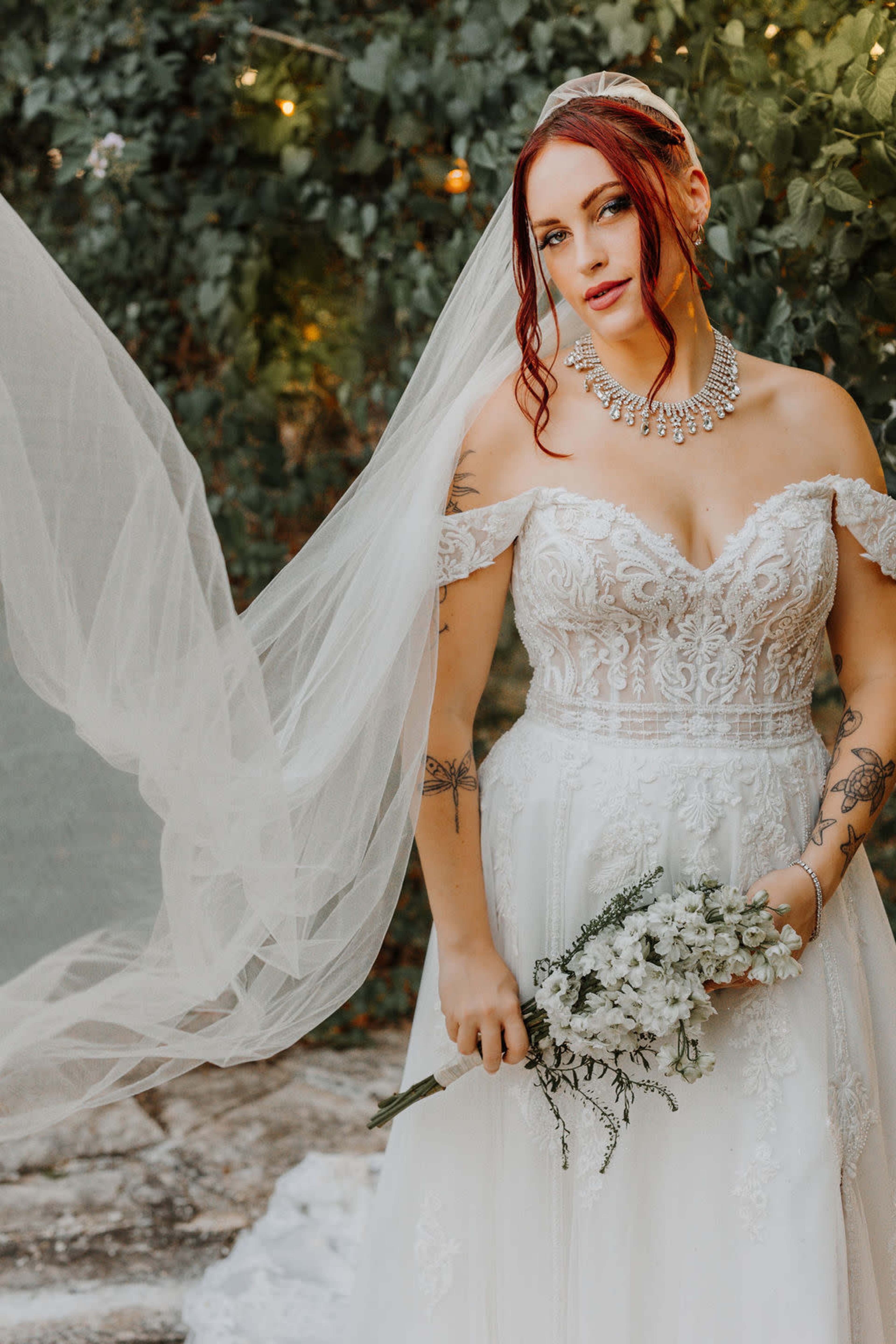 A bride wearing a detailed white gown and veil stands outdoors, holding a bouquet of flowers against a backdrop of greenery.