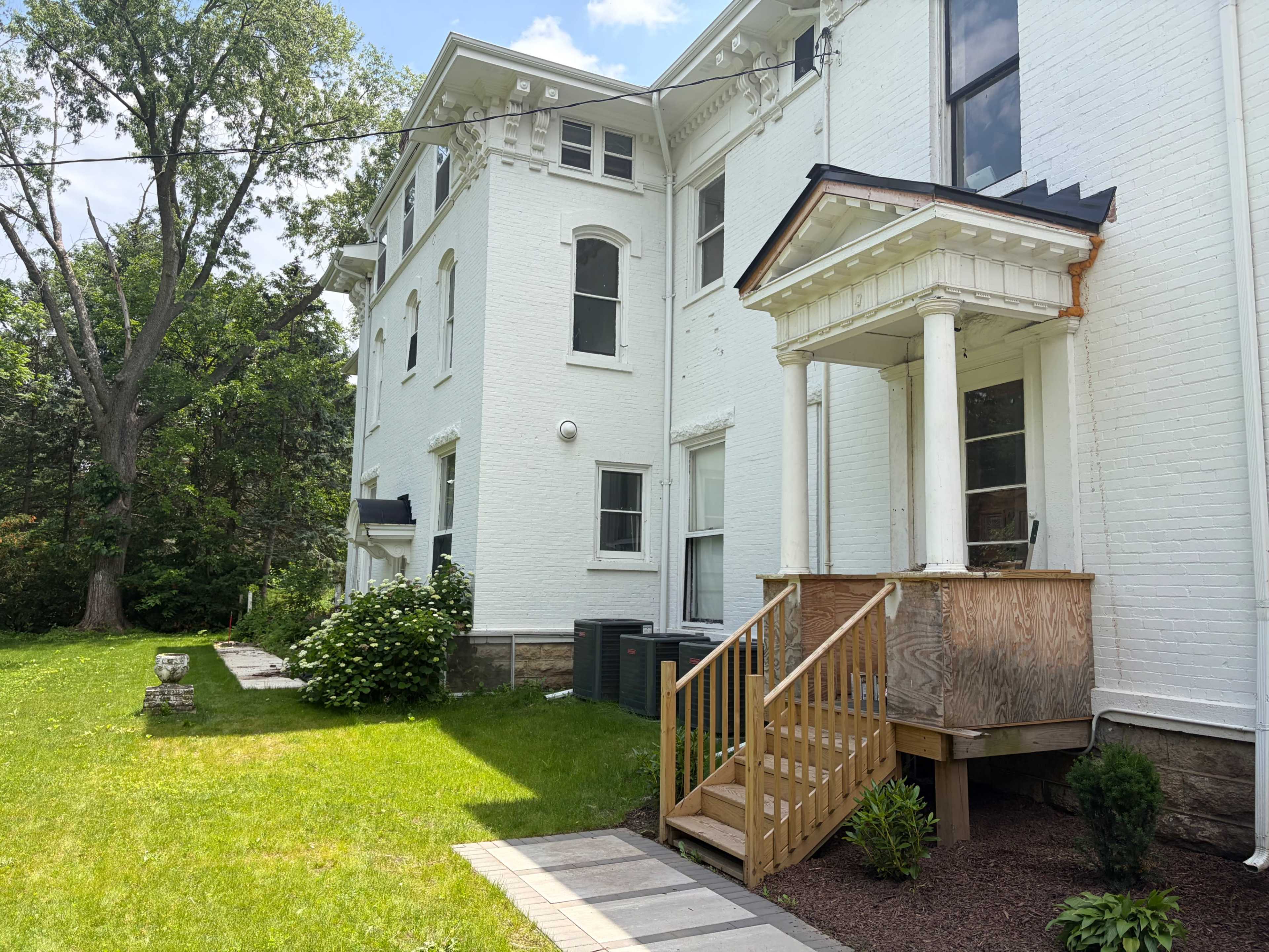 The image shows a white brick building with a wooden porch and steps, surrounded by green grass and trees.