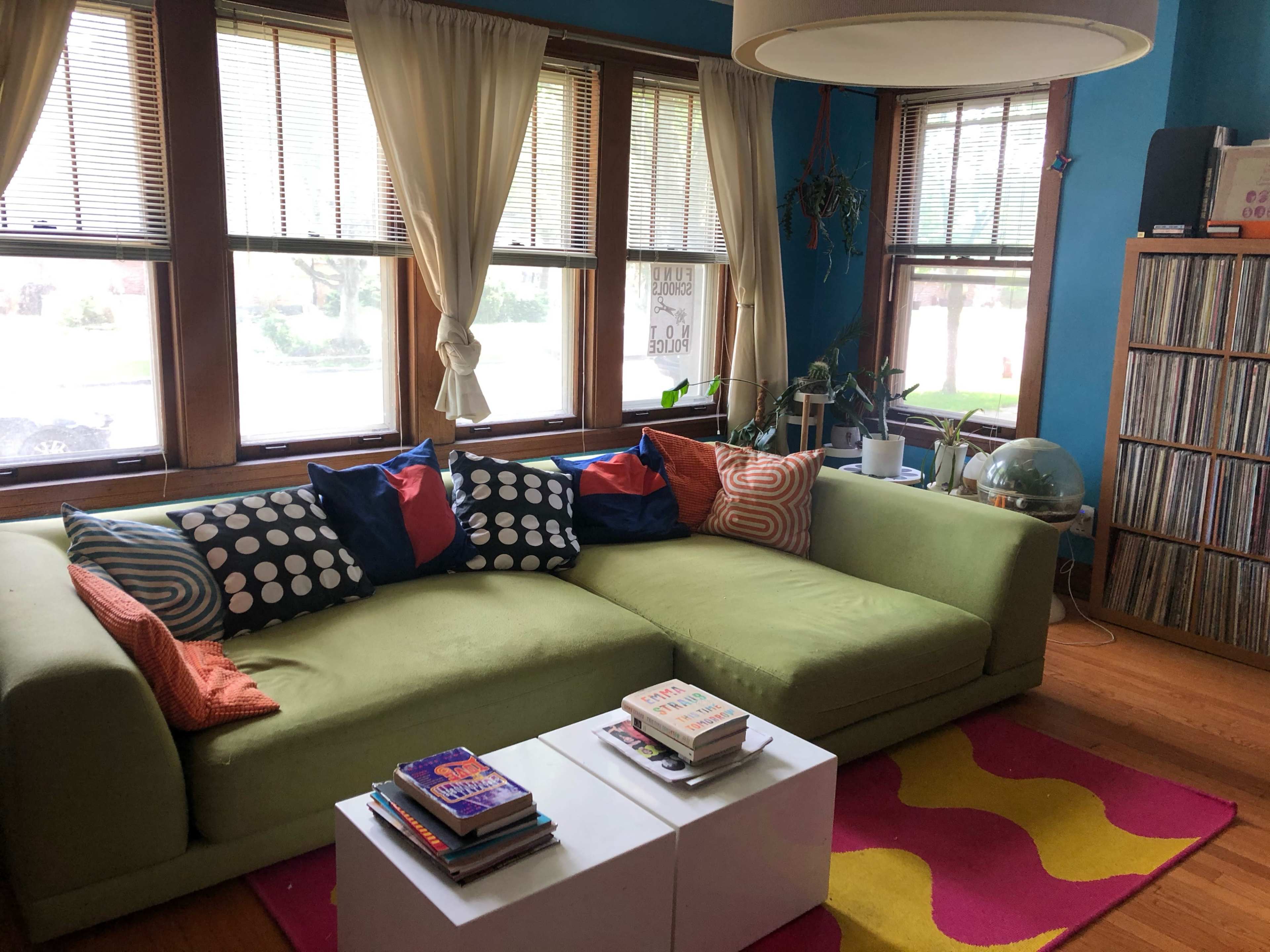A green sectional sofa with colorful throw pillows is positioned against a wall of windows, accompanied by a coffee table and a collection of books on the floor in a brightly lit room.