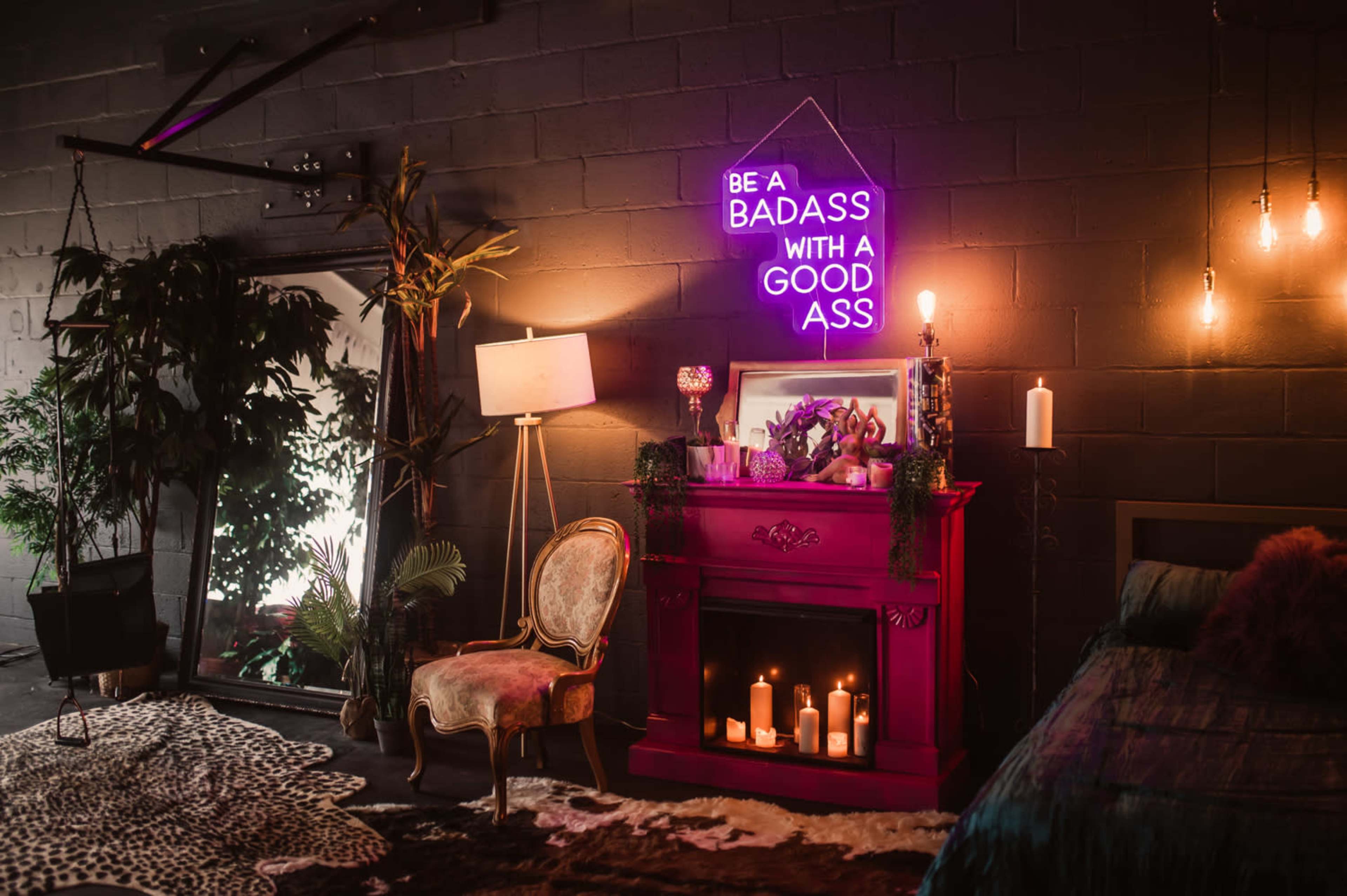 A dimly lit room featuring a bright pink fireplace adorned with candles, a neon sign, a vintage chair, and decorative plants.
