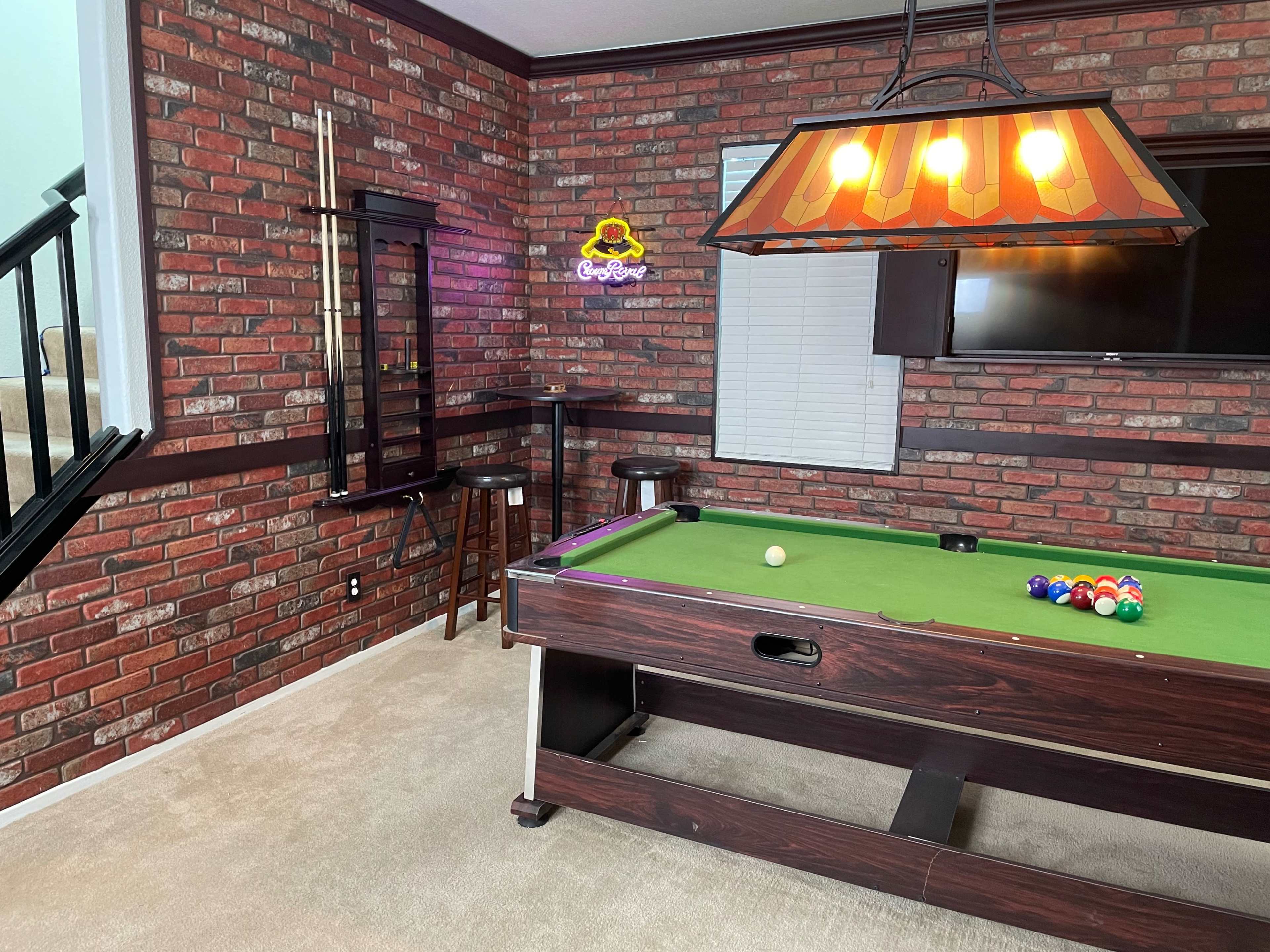 A game room featuring a pool table against a brick-patterned wall, with a bar area and a mounted television.