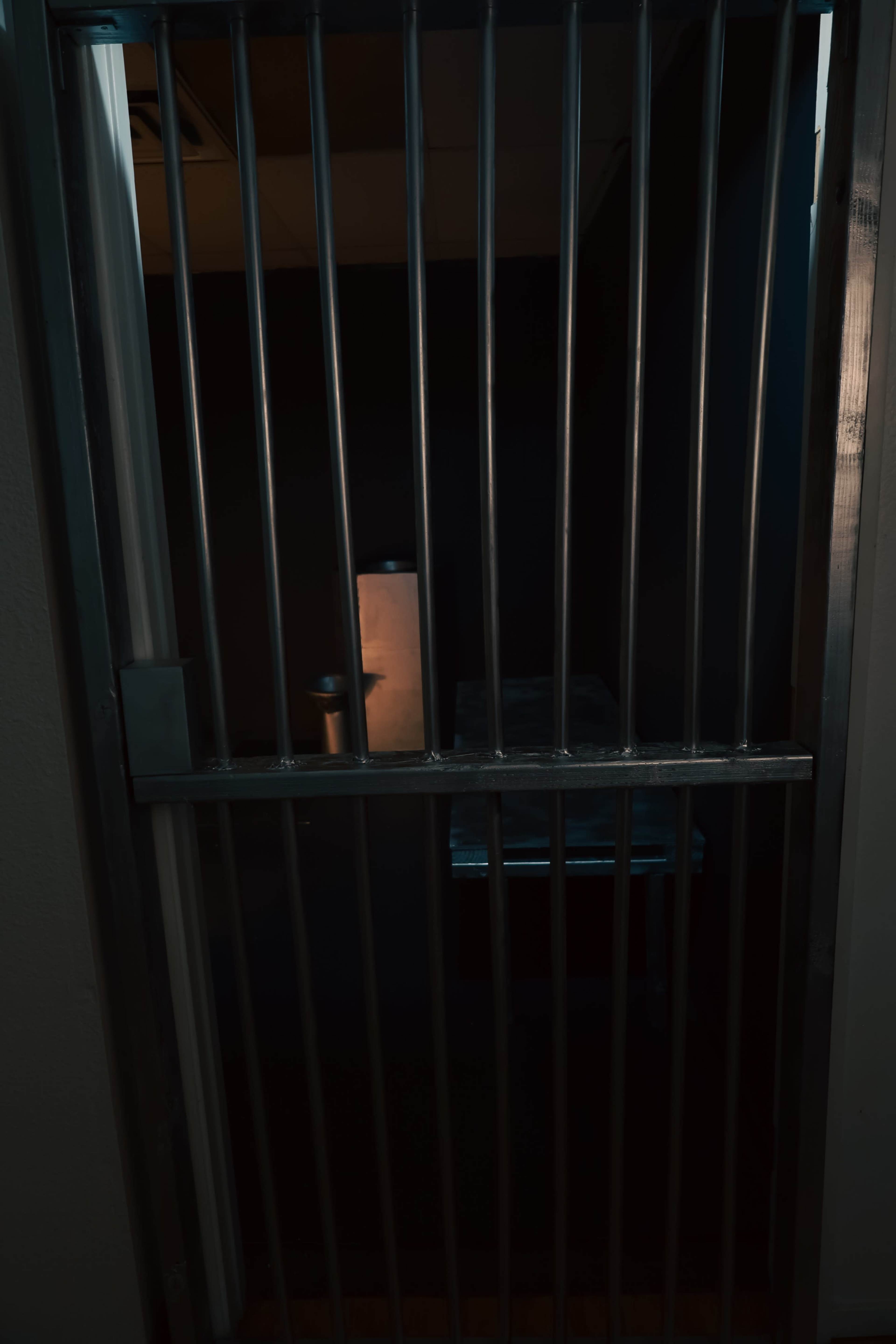 The image shows the barred entrance of a dimly lit jail cell, with a small table and a light source visible inside.