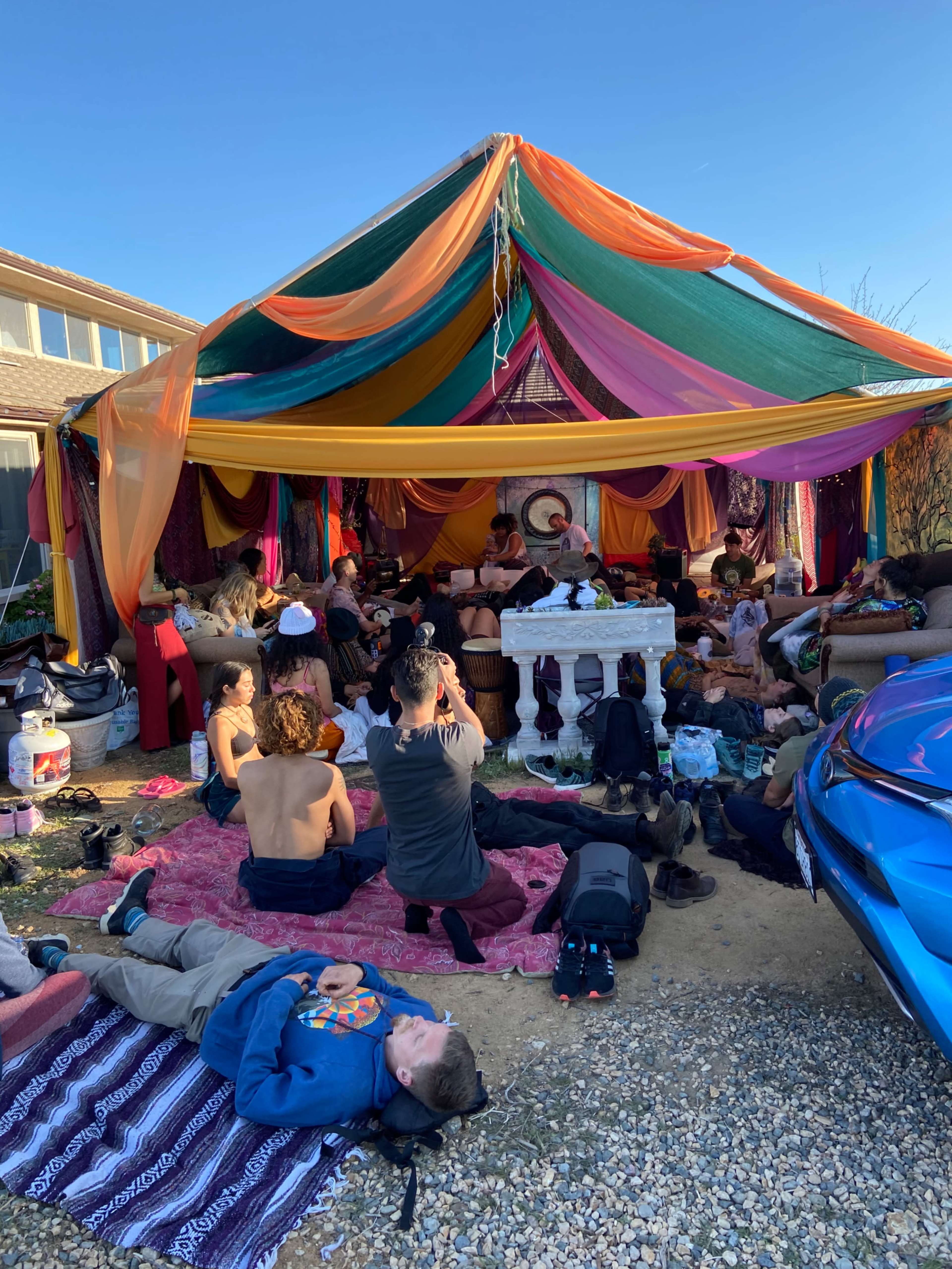 A large colorful canopy covers a gathering of people sitting on the ground and lounging in a casual outdoor setting.