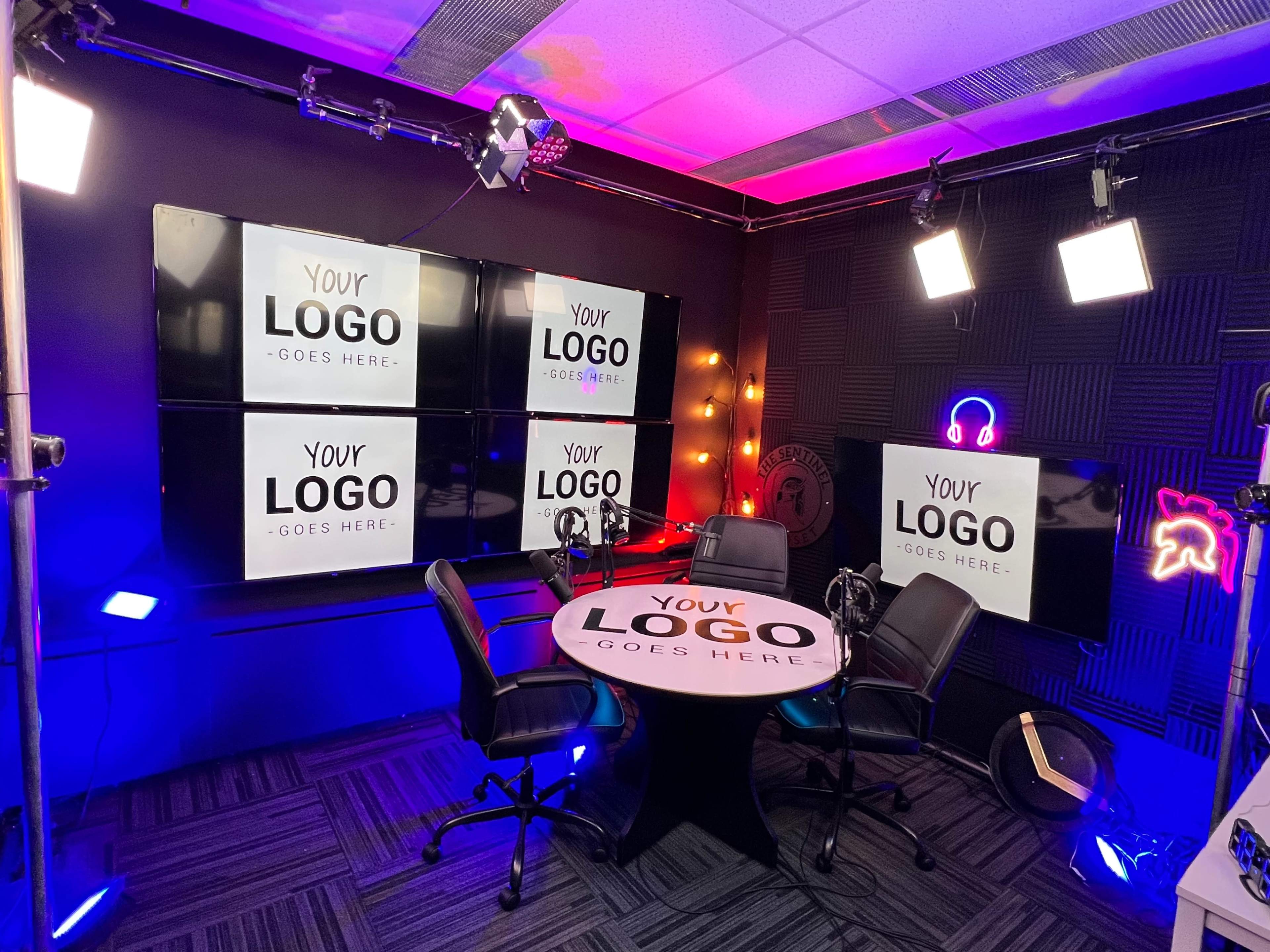 The image shows a modern studio setup for video production, featuring a round table with two chairs, multiple large screens displaying the text "YOUR LOGO GOES HERE," and various lighting fixtures.