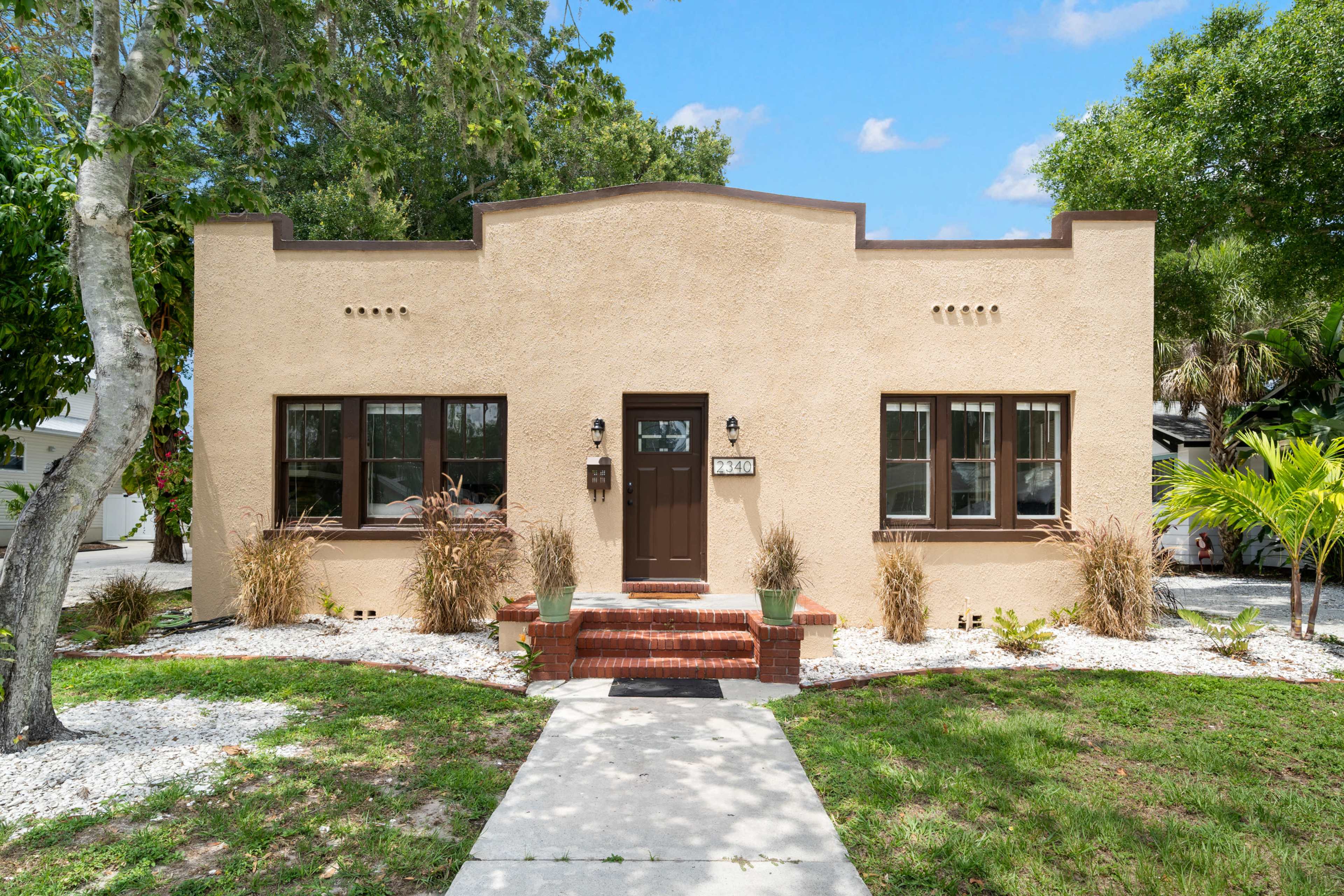 A single-story, stucco house with a brown door and windows, surrounded by landscaped grass and decorative plants.