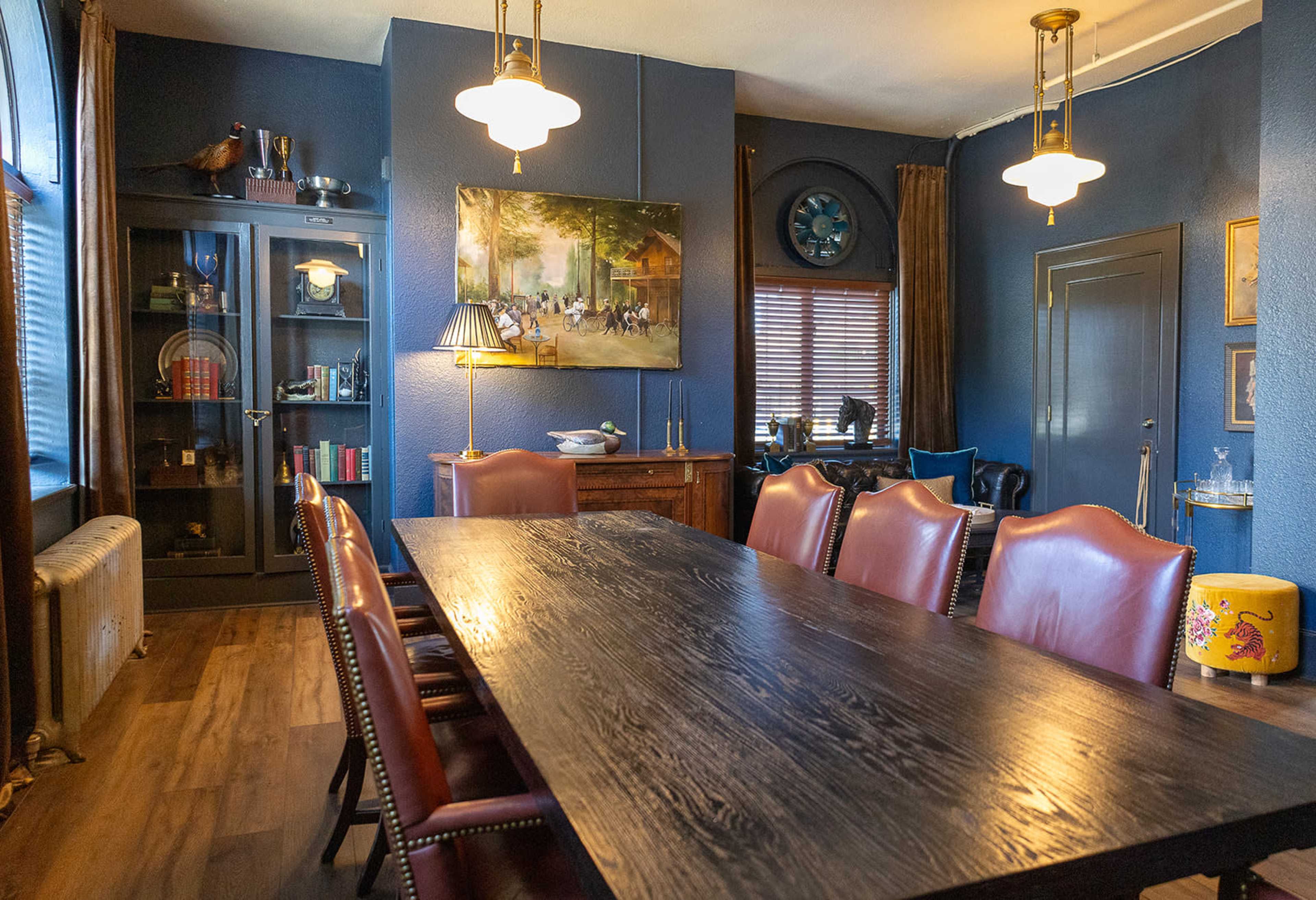 The image shows a dining room with a long wooden table surrounded by leather chairs, accented by a large painting and shelves filled with decorative items.