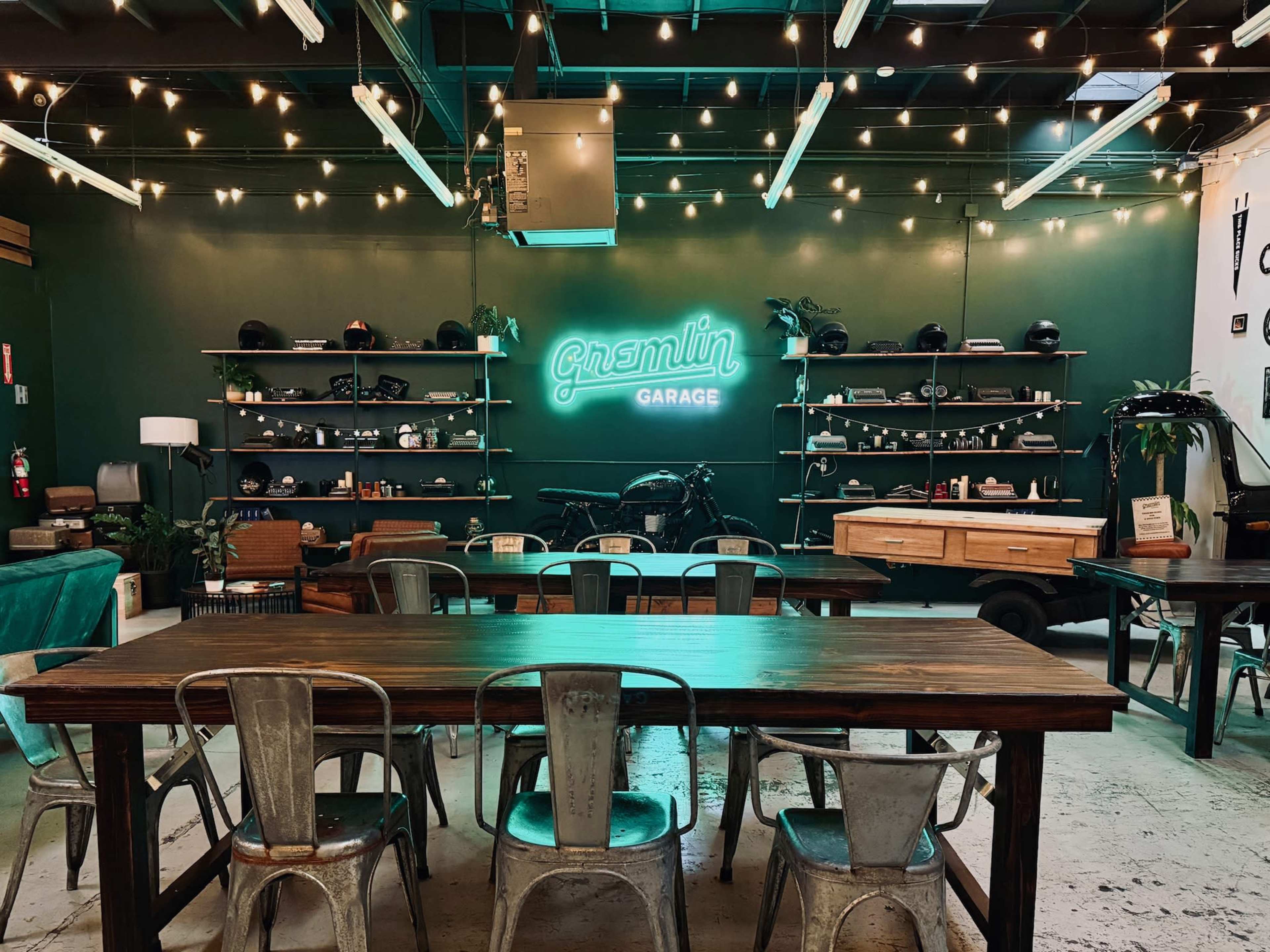 The image shows a stylish garage interior featuring a long wooden table surrounded by metal chairs, neon signage, and shelves displaying various items against a dark green backdrop.