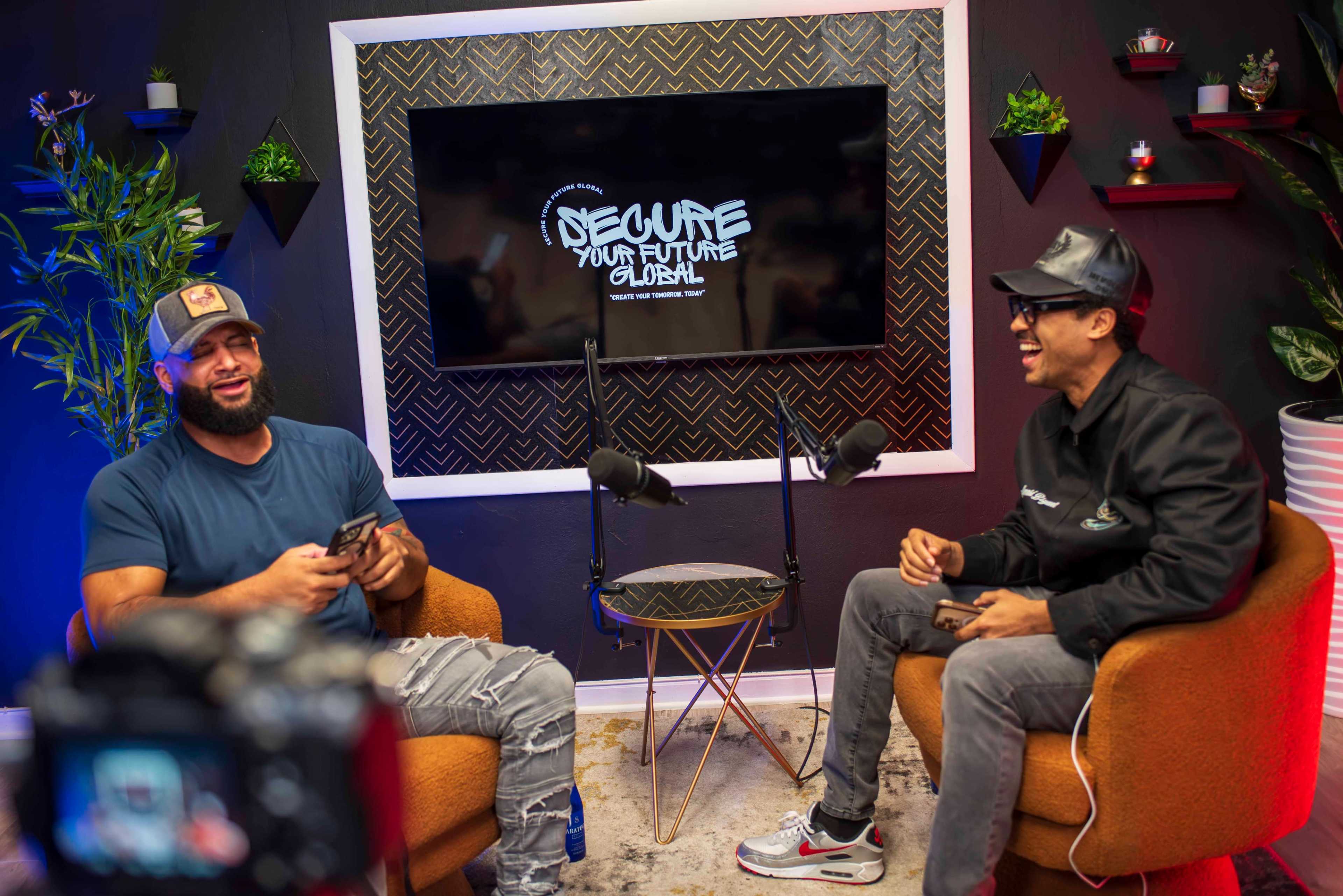 Two men are seated in a studio setting with microphones, laughing and engaging in conversation, framed by a screen that reads "SECURE YOUR FUTURE GLOBAL."