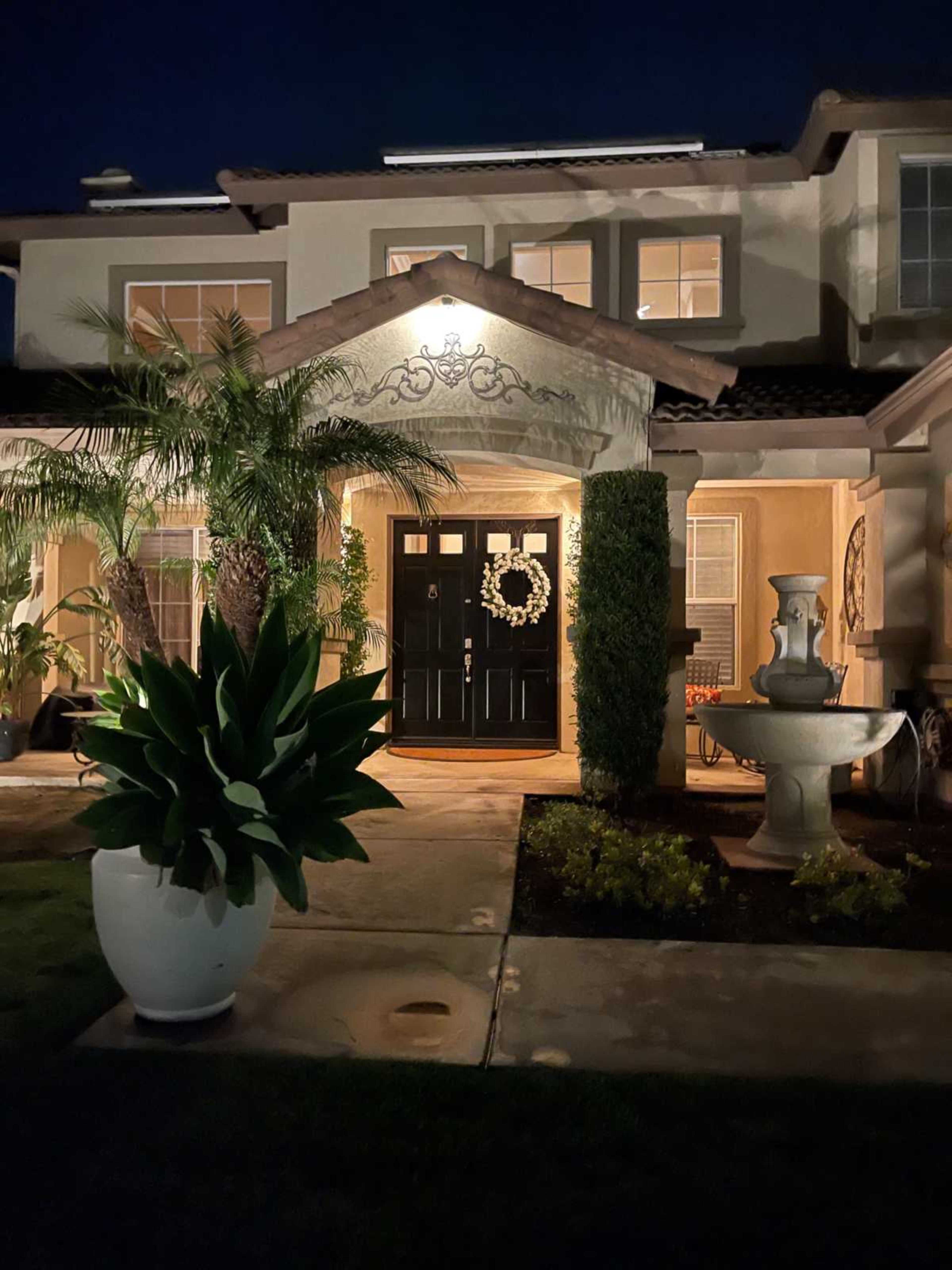 The image shows a well-lit entryway of a home featuring a dark front door, decorative wreath, and a fountain surrounded by lush greenery.