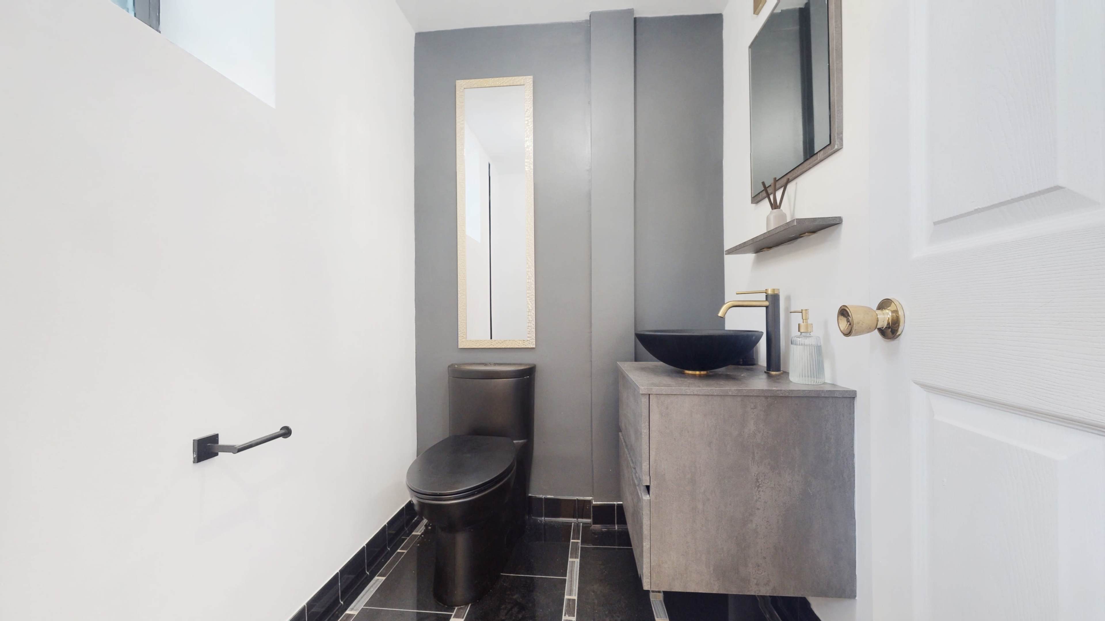 A modern bathroom featuring a black toilet, a dark countertop basin, a mirror, and gray and white walls with black tiled flooring.