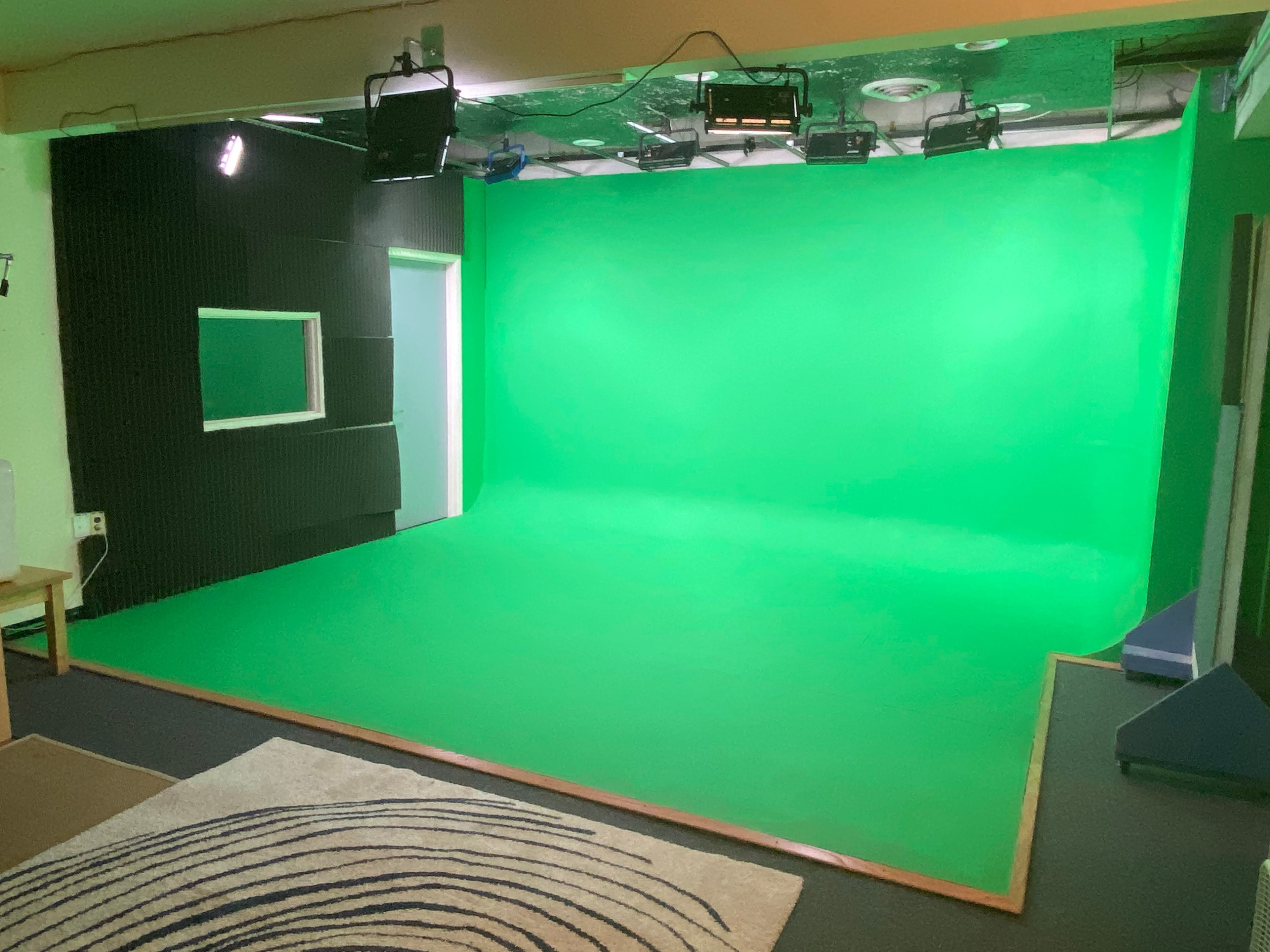A green screen studio with a flat green backdrop, track lighting, and a small window in the black acoustic wall.