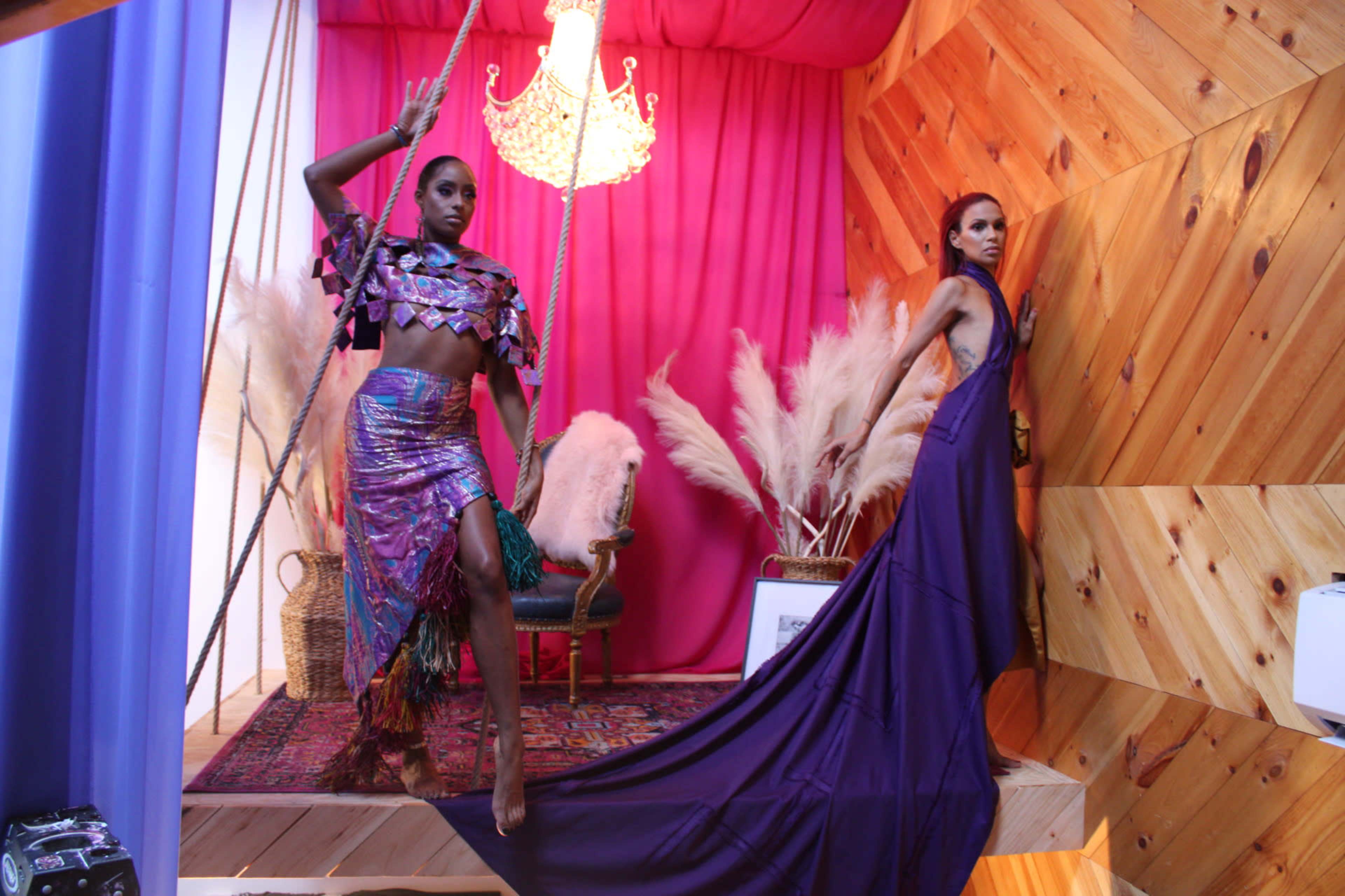 Two models pose in a colorful set featuring a swing and a chandelier, with vibrant fabrics and decorative pampas grass in the background.