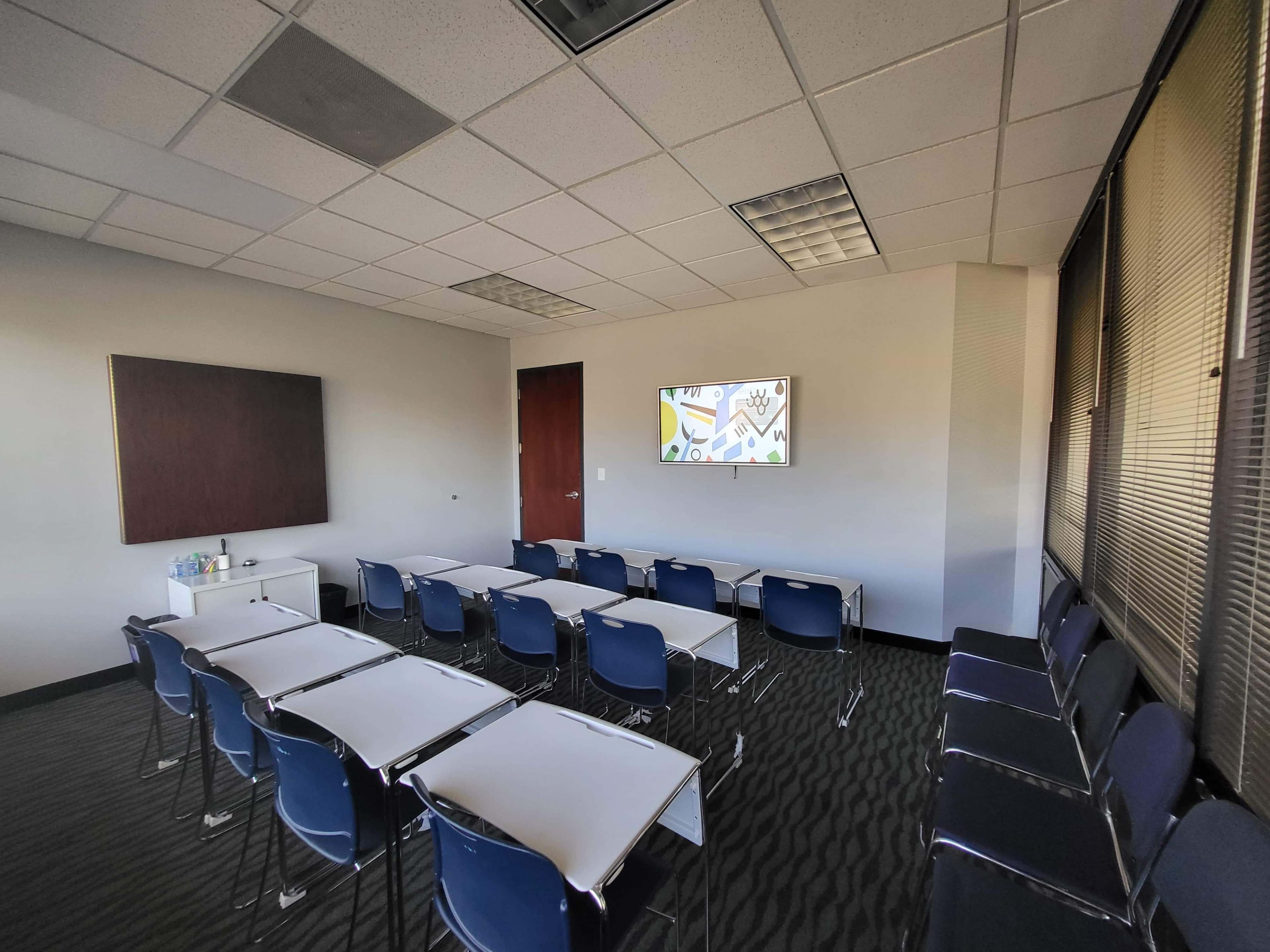 A modern classroom with rows of blue chairs and tables facing a wall-mounted screen, along with a large window.