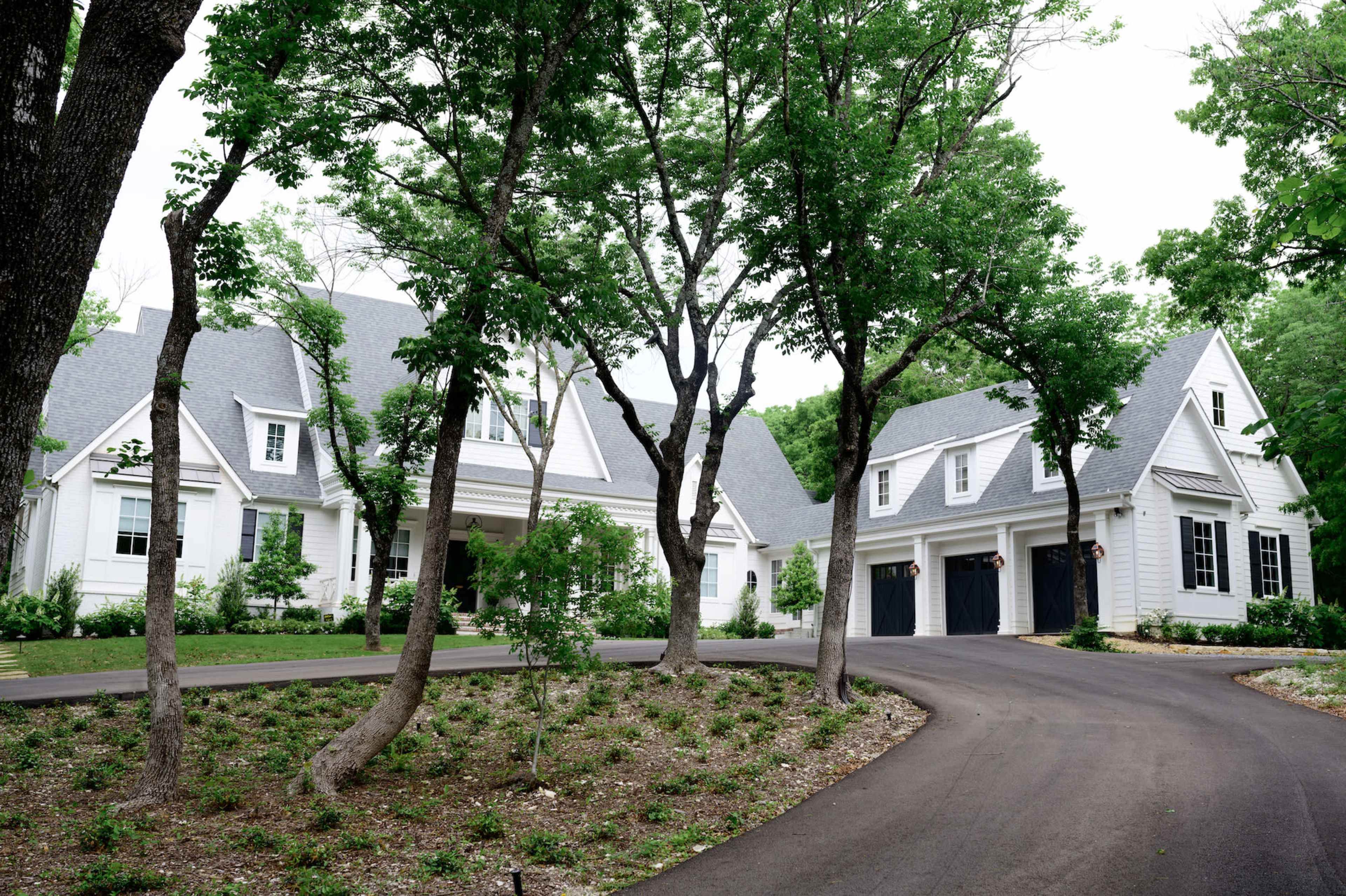 A large white house with a gray roof is set among trees, with a circular driveway leading to a three-car garage.