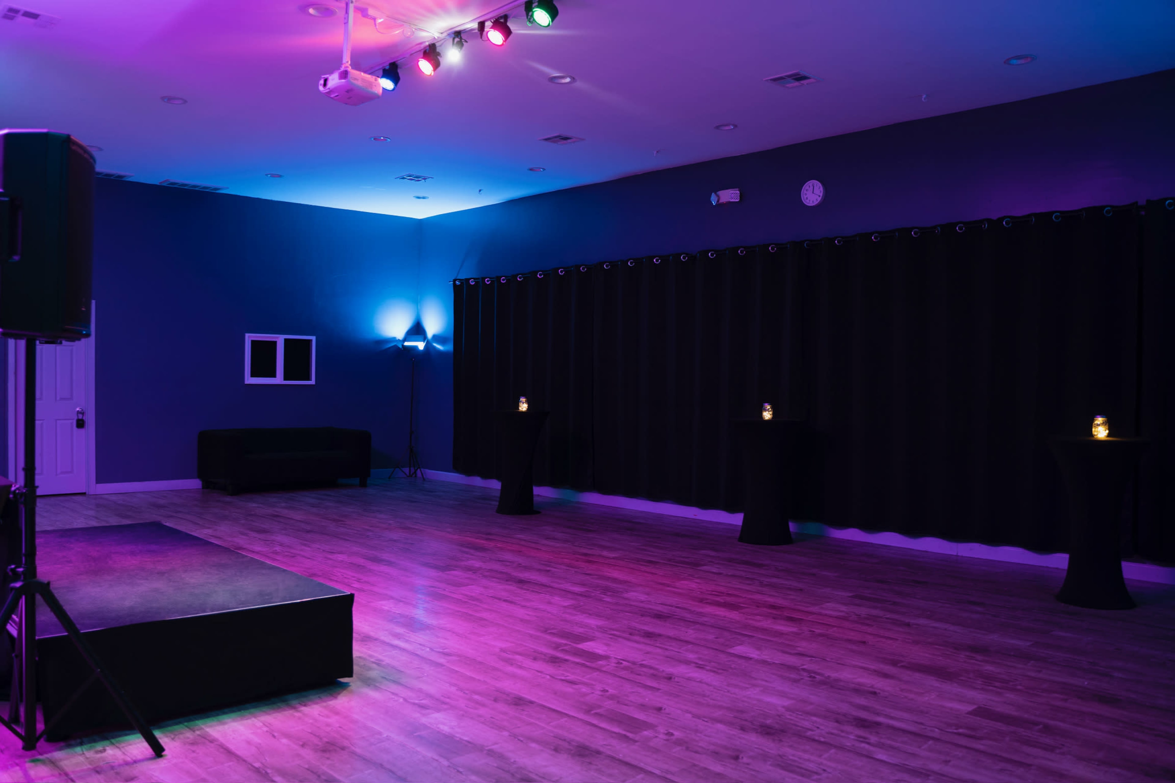 A dimly lit event space features a small stage, black curtains, and several illuminated pedestal tables.