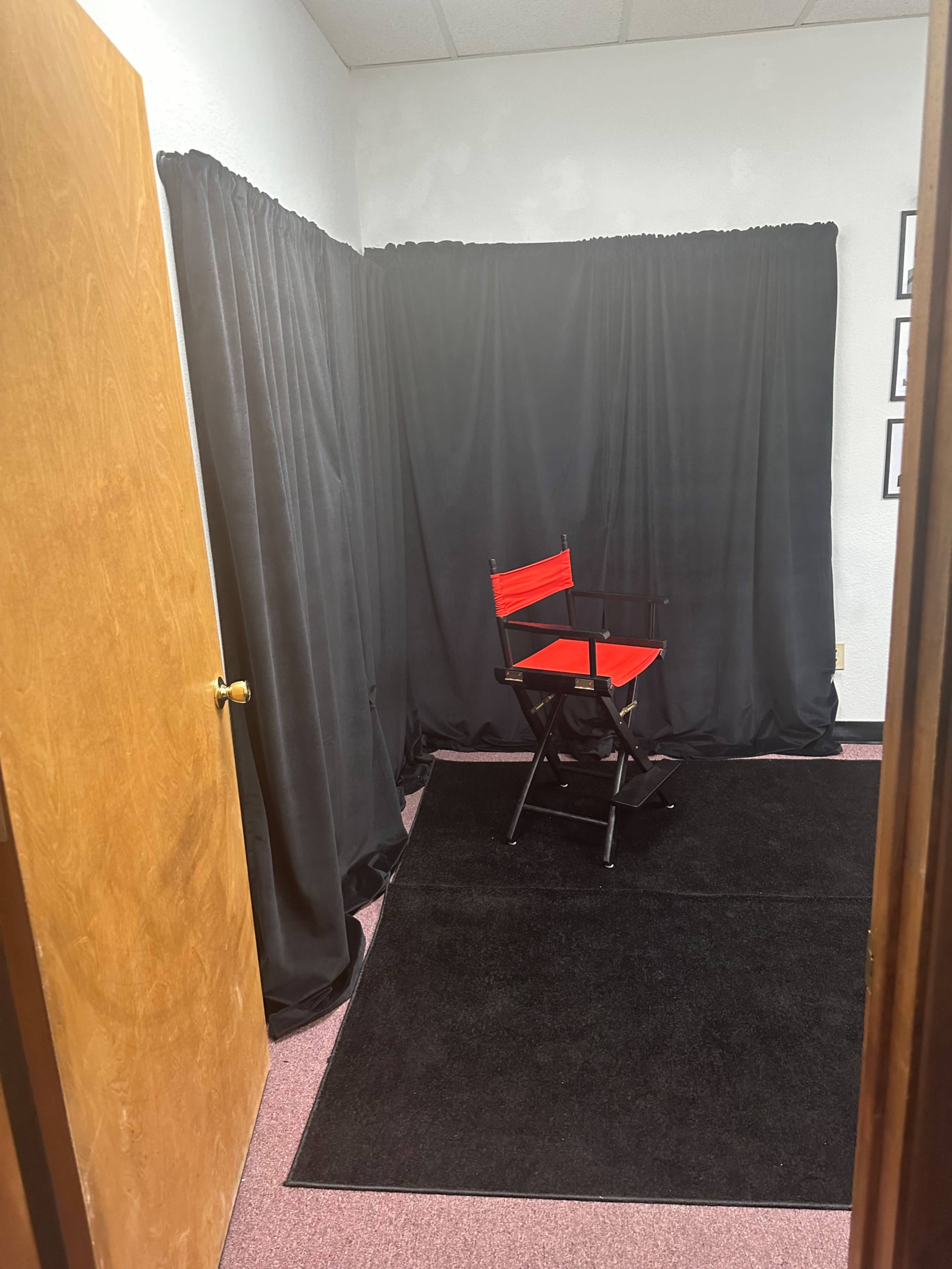 A small room with a black curtain backdrop, a red director's chair, and a dark carpet, with a wooden door partially open.