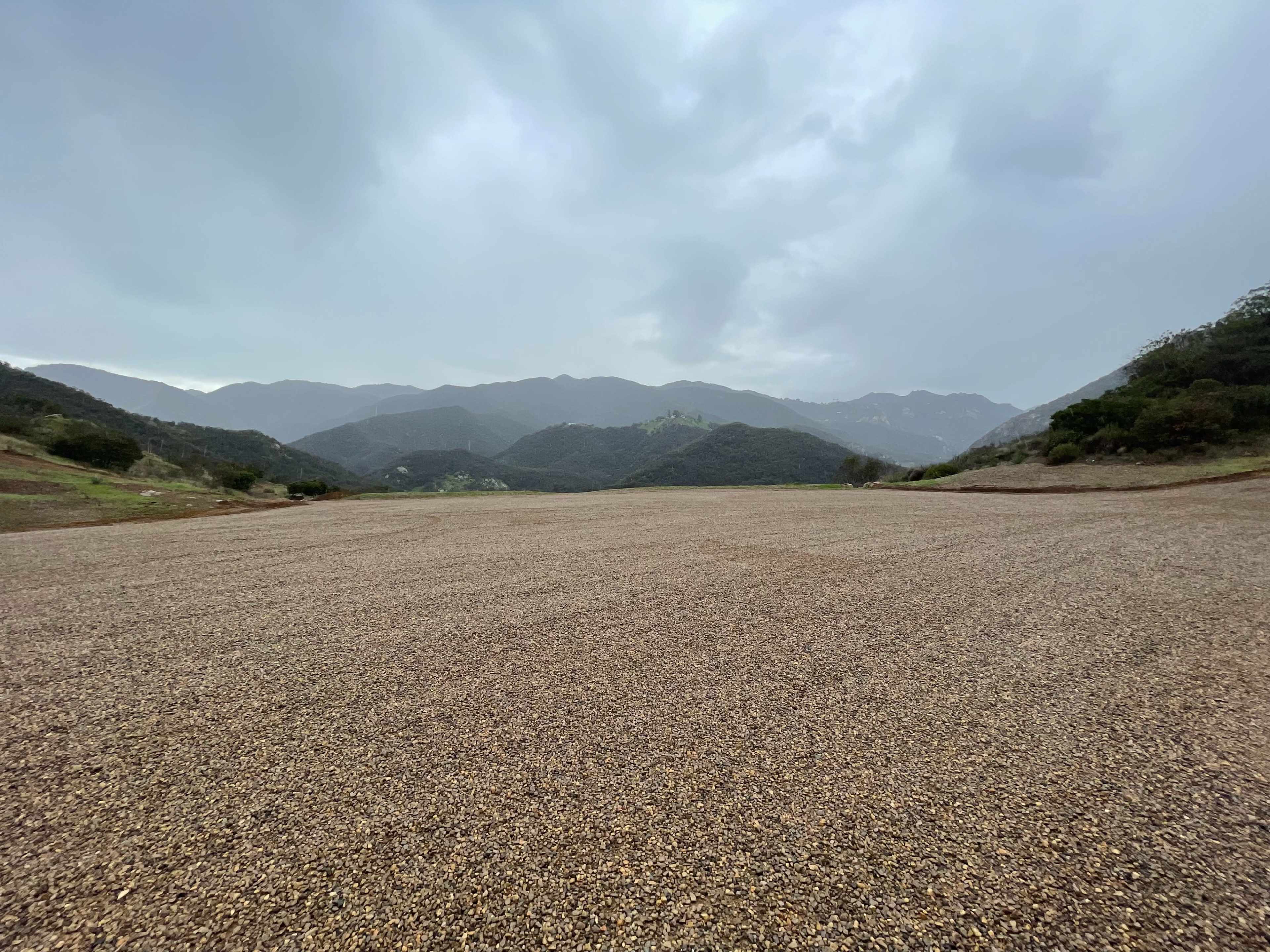 A gravel expanse stretches across the foreground, bordered by hills and mountains under a cloudy sky.