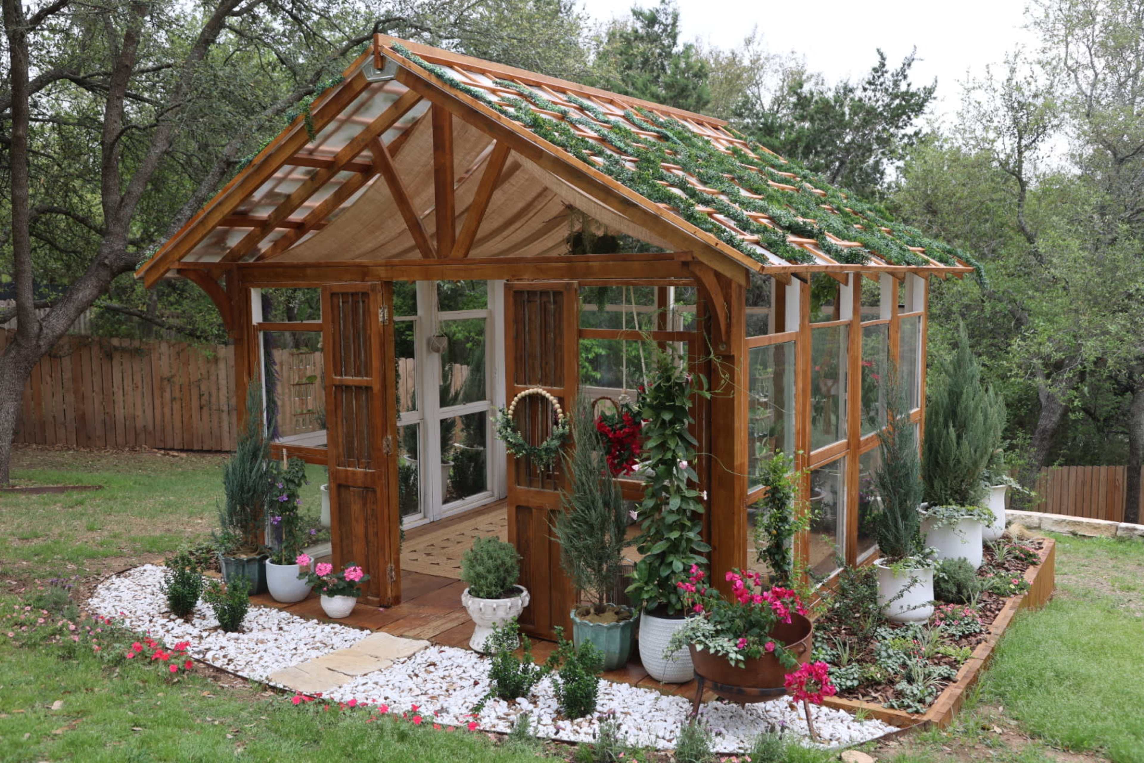A wooden greenhouse with large glass panels features a sloped roof adorned with greenery, surrounded by garden beds and potted plants.