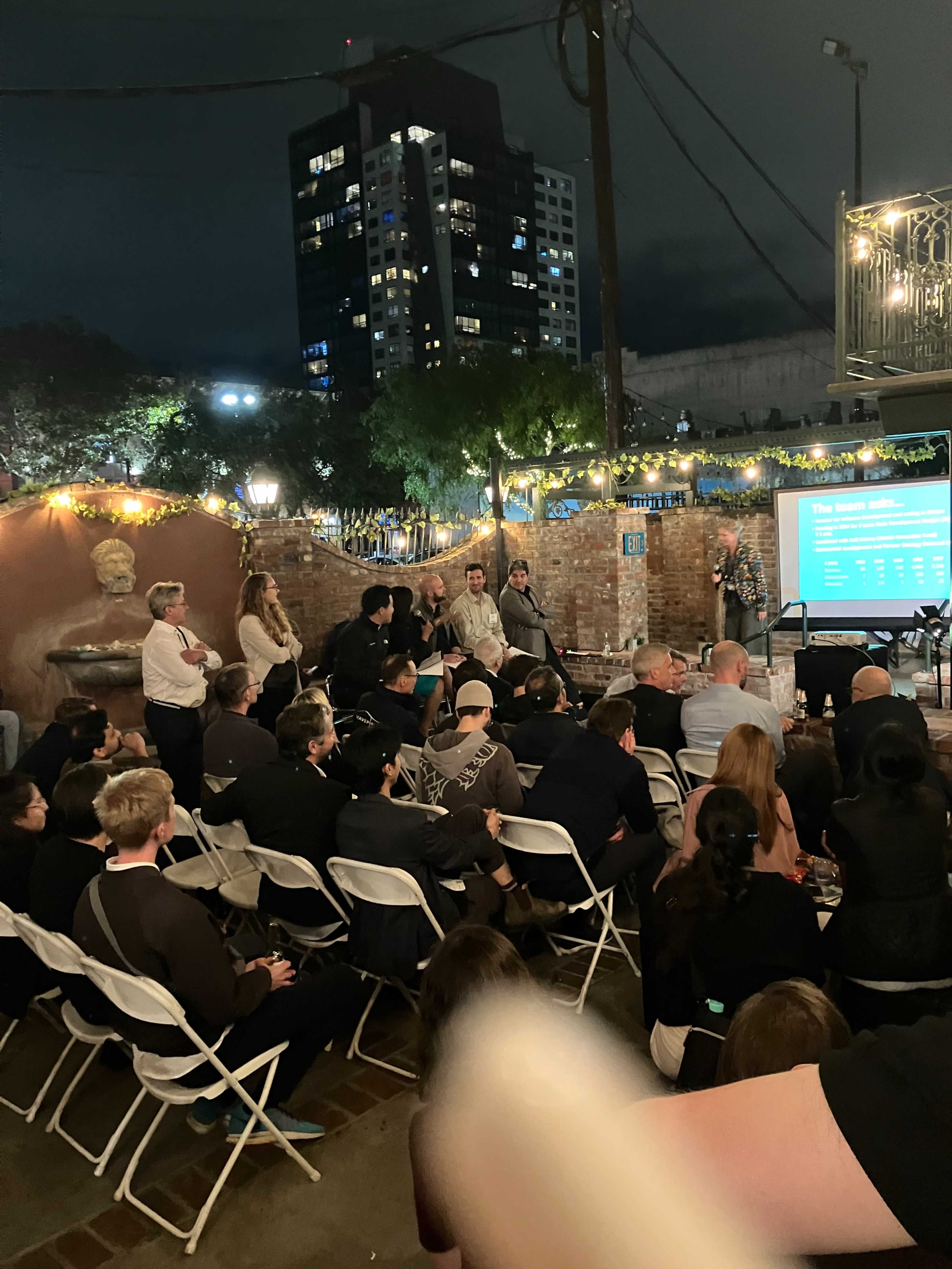 A crowd of people sits in folding chairs at an outdoor event illuminated by string lights, with a building featuring large windows in the background.