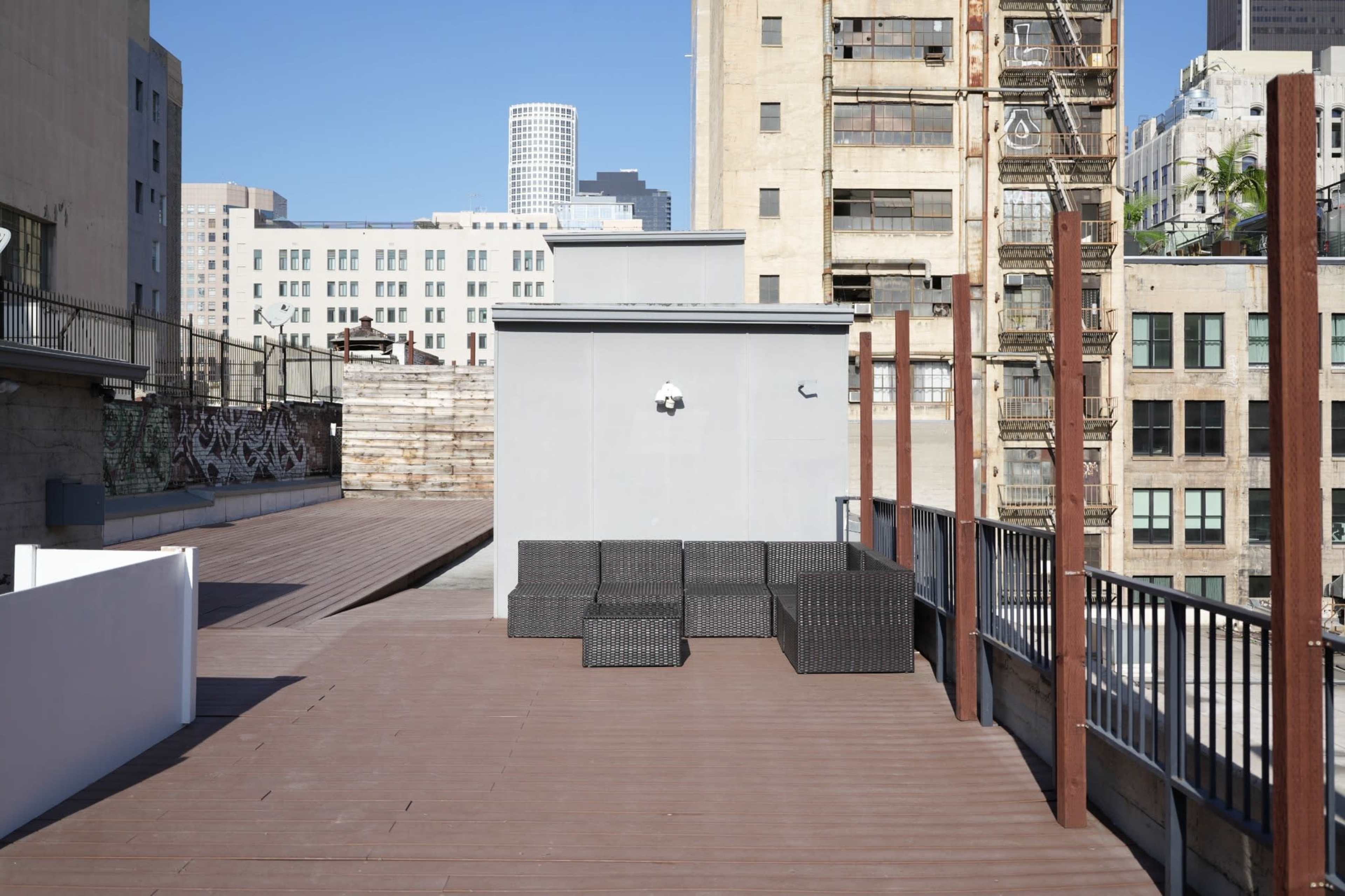 The image shows a flat rooftop deck with wooden flooring, featuring several dark wicker seating arrangements and a backdrop of urban buildings.