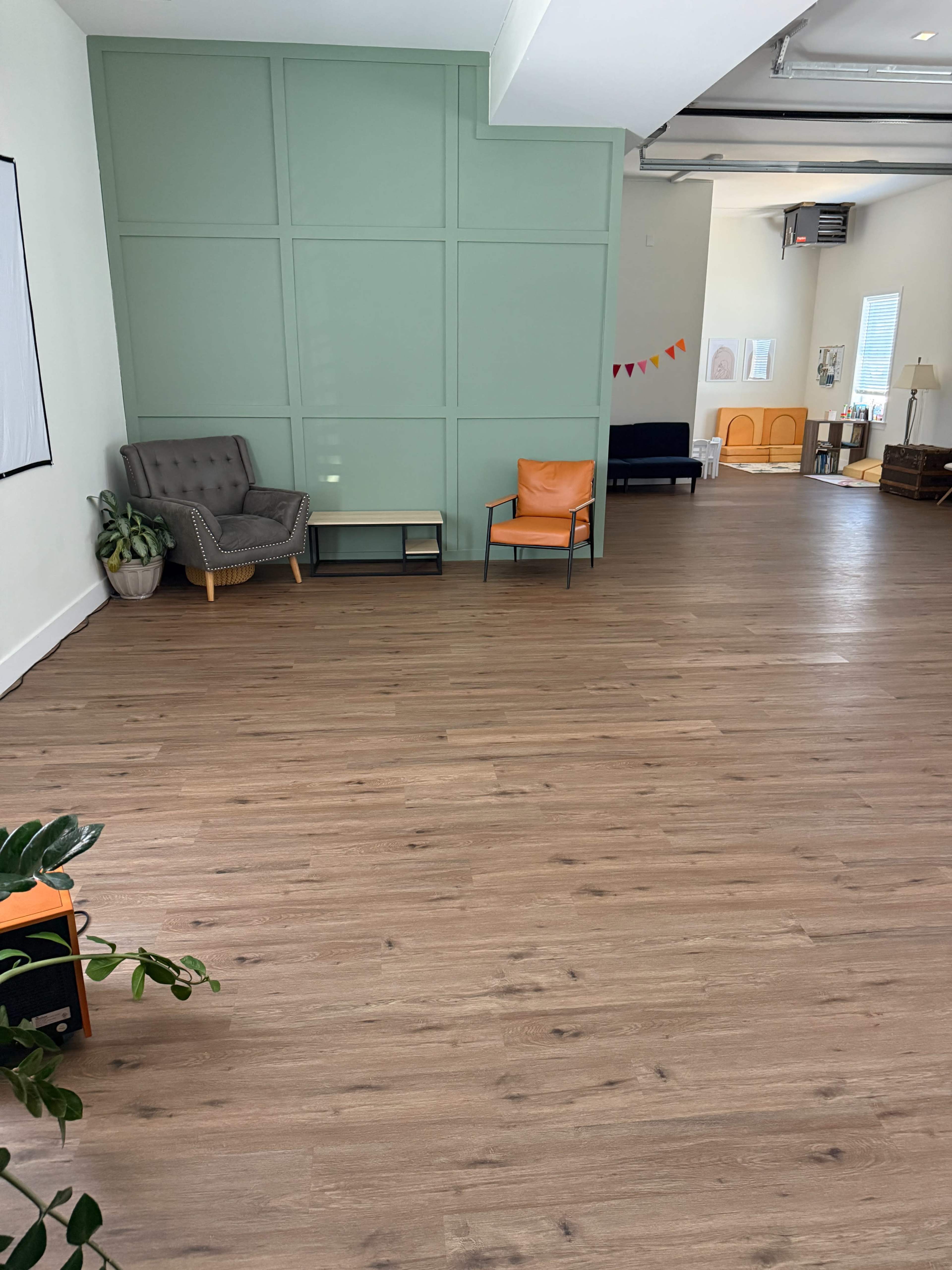 The image shows a spacious, light-filled room with wooden flooring, featuring a green accent wall, two chairs in different colors, and minimal furniture.