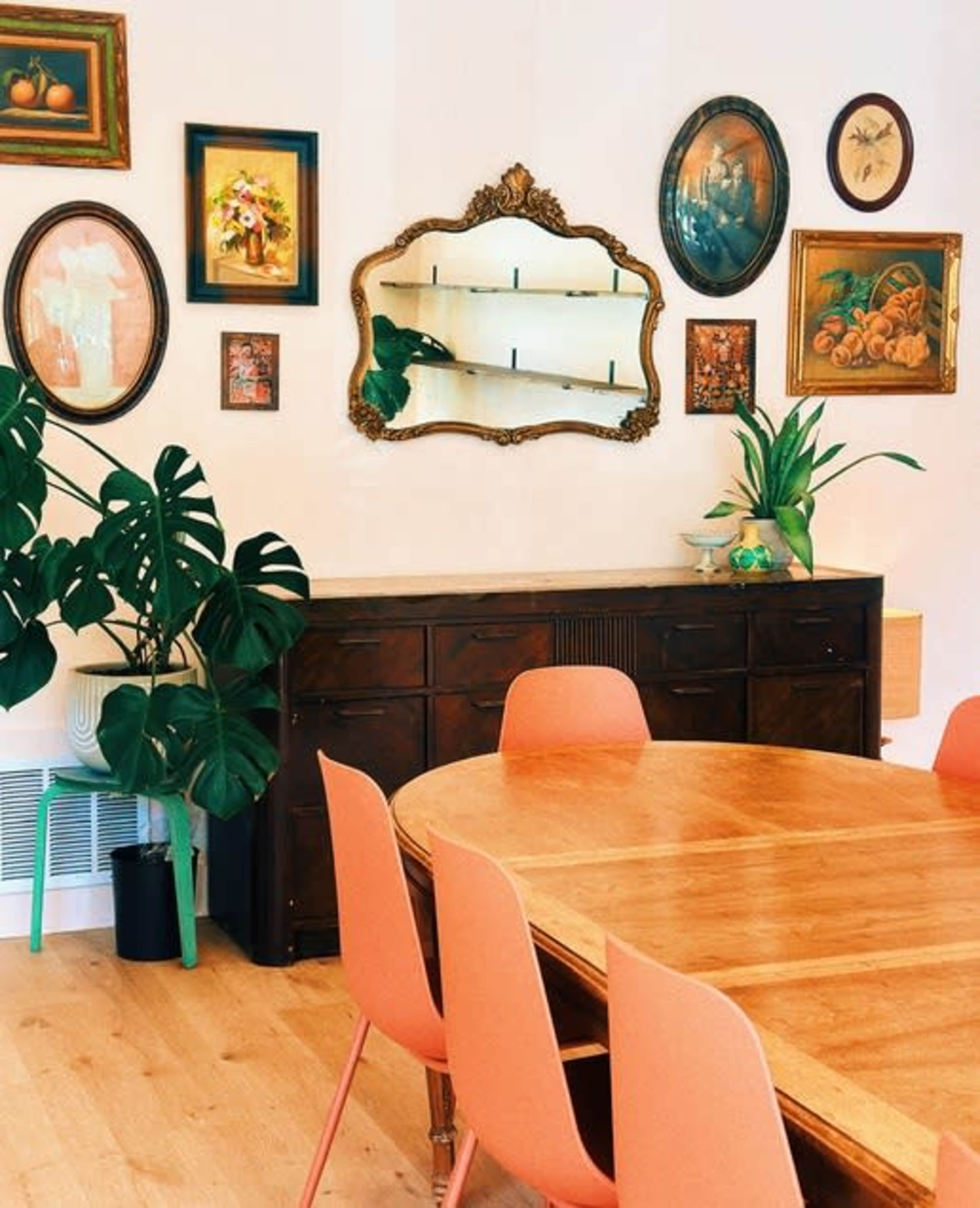 The image features a dining area with a round wooden table surrounded by pink chairs, a dark wooden sideboard, and various framed artworks on the wall above it.