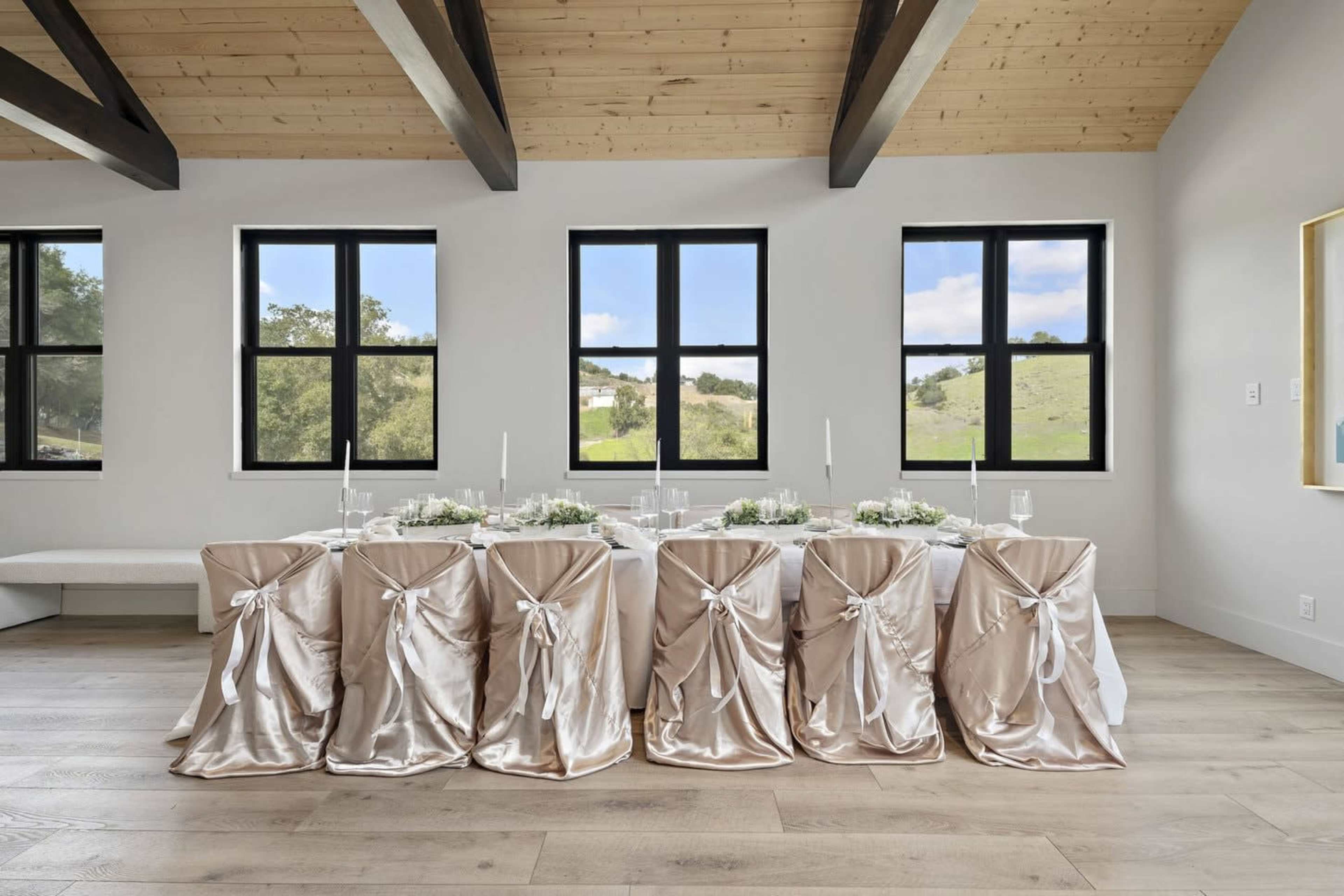 A long dining table is set with elegant tableware, surrounded by chairs draped in satin fabric, against a backdrop of large windows overlooking a green landscape.