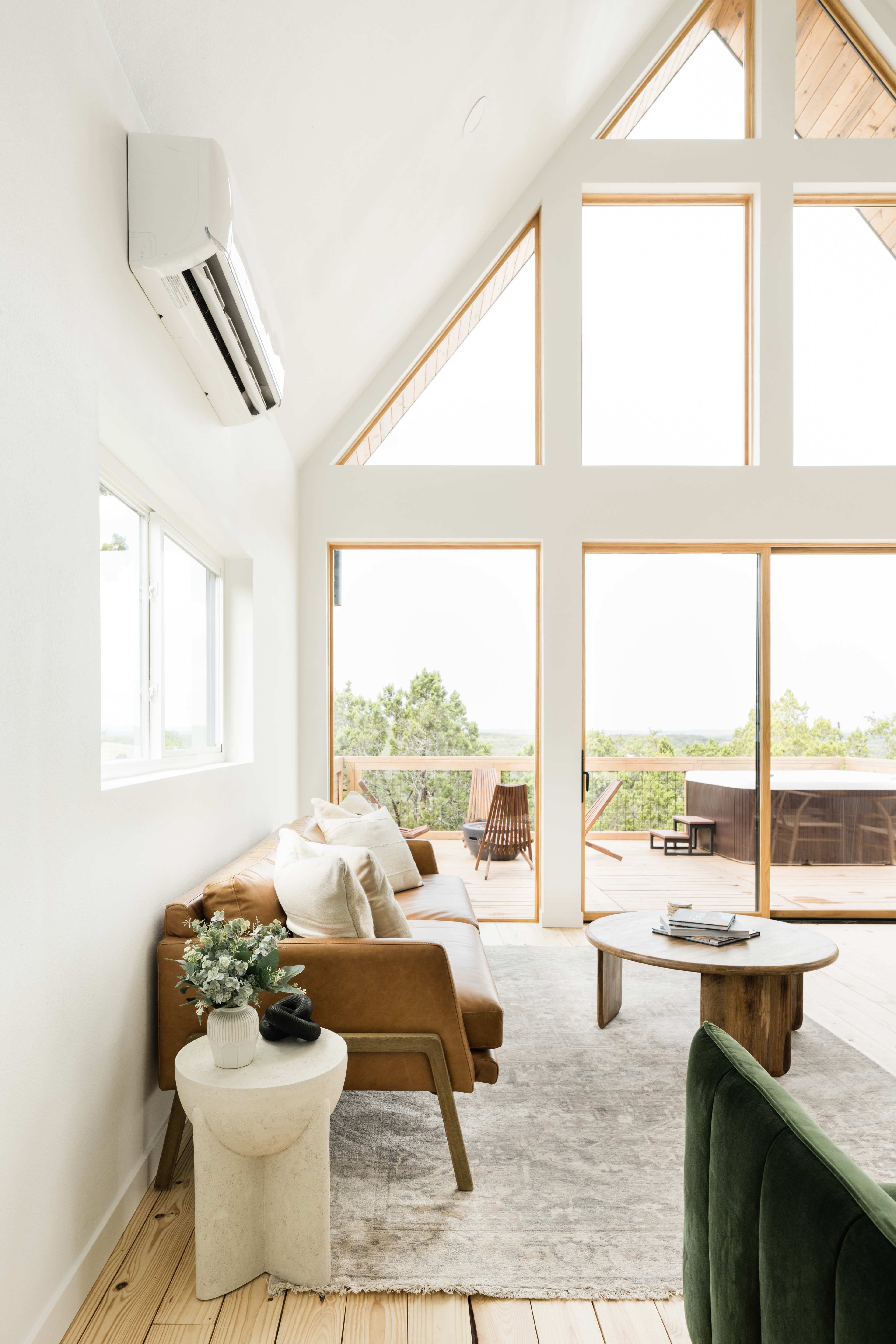 The interior of a modern living room features large windows, a leather sofa, a wooden coffee table, and a view of an outdoor deck.