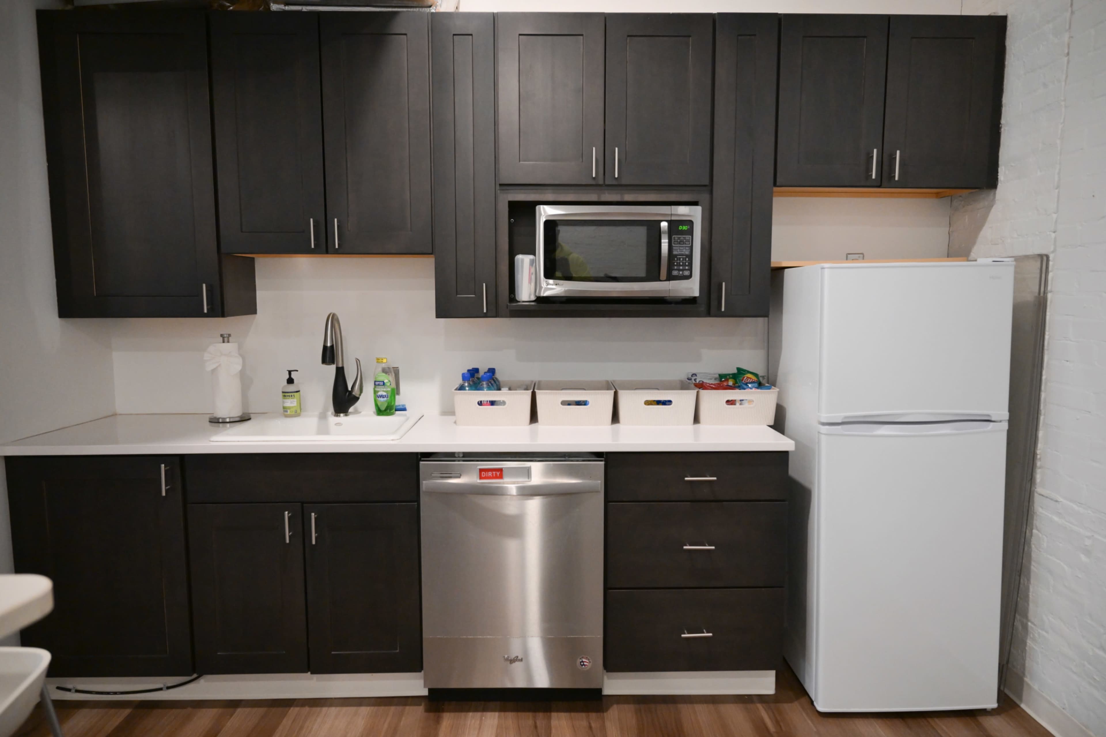 The image shows a modern kitchen with dark cabinetry, a stainless steel dishwasher, a microwave above the countertop, and a white refrigerator.