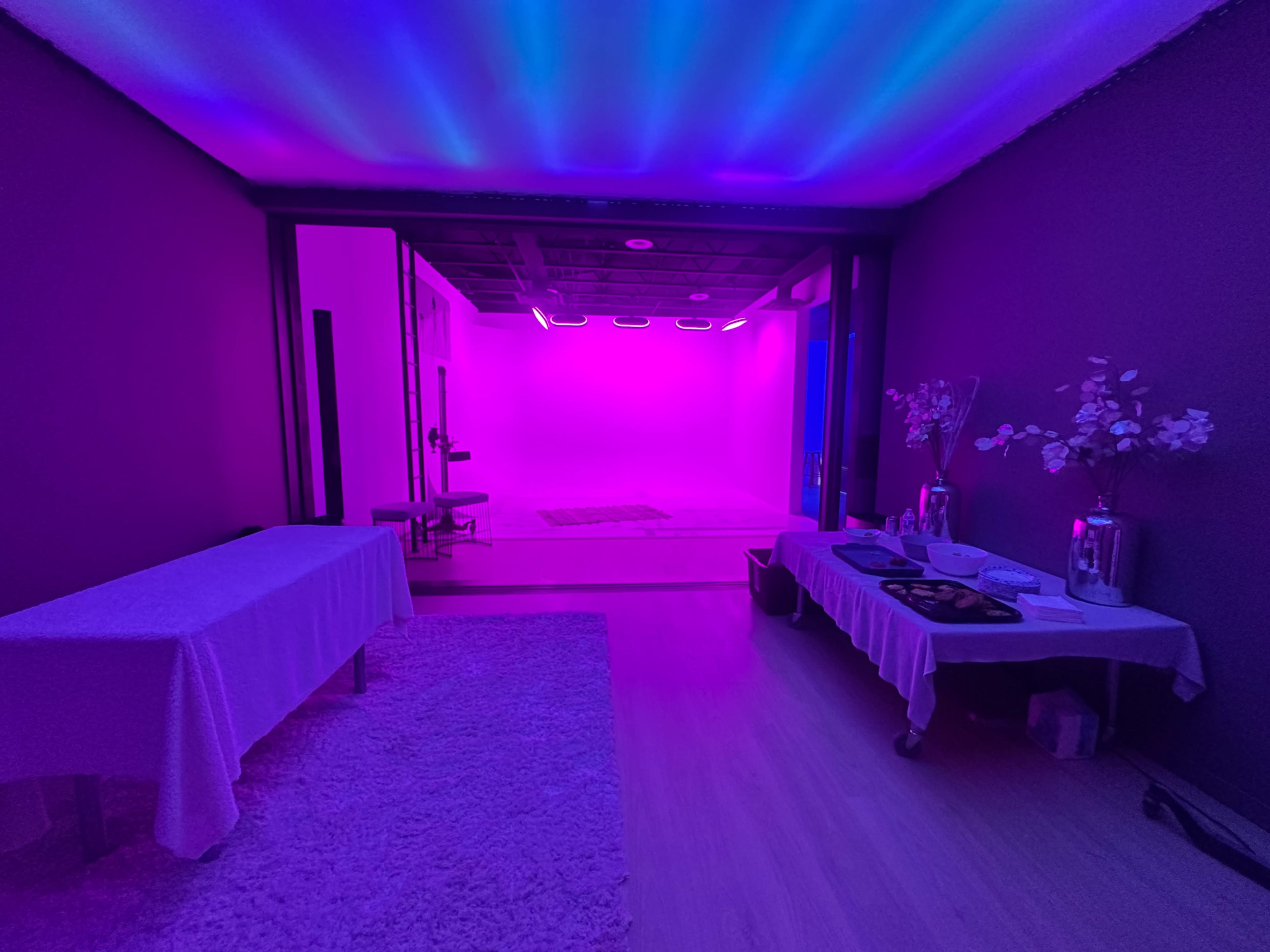 A room features two tables with white tablecloths and snacks, leading to a space illuminated with vibrant pink and purple lighting.