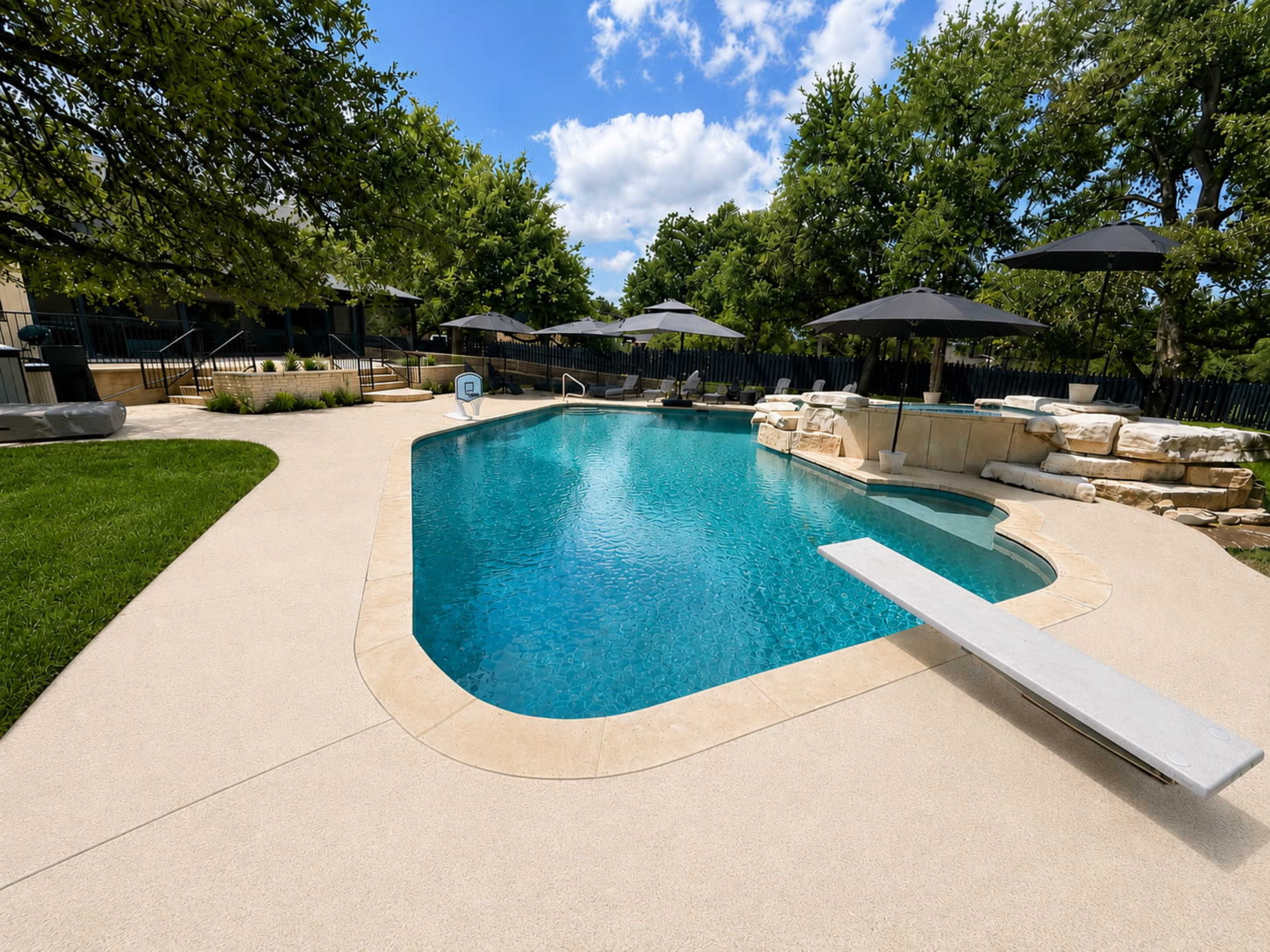 A swimming pool with a diving board is surrounded by trees and shaded areas with umbrellas.