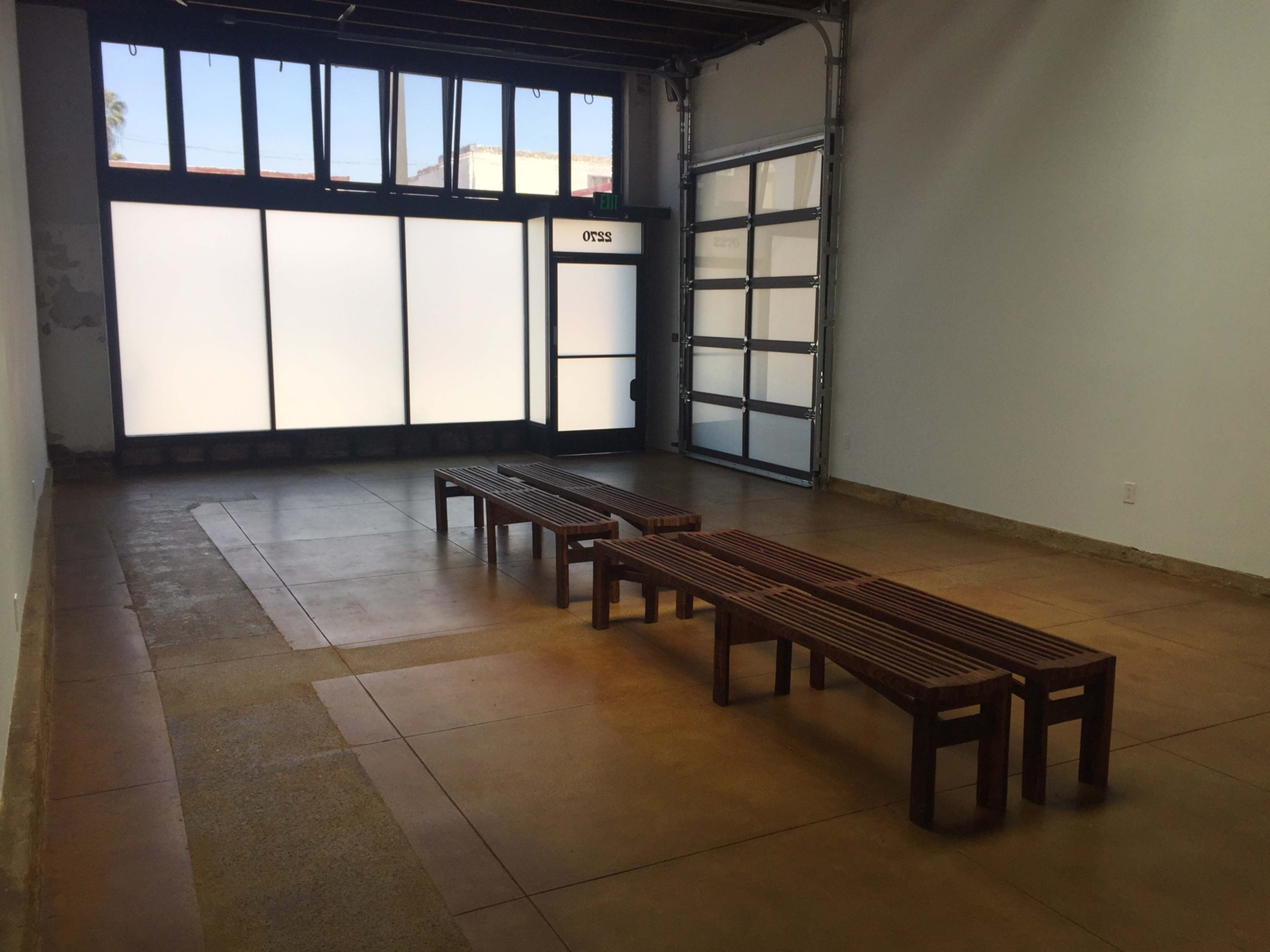 The image shows an empty room with concrete flooring, two wooden benches, and large windows that allow natural light to enter.