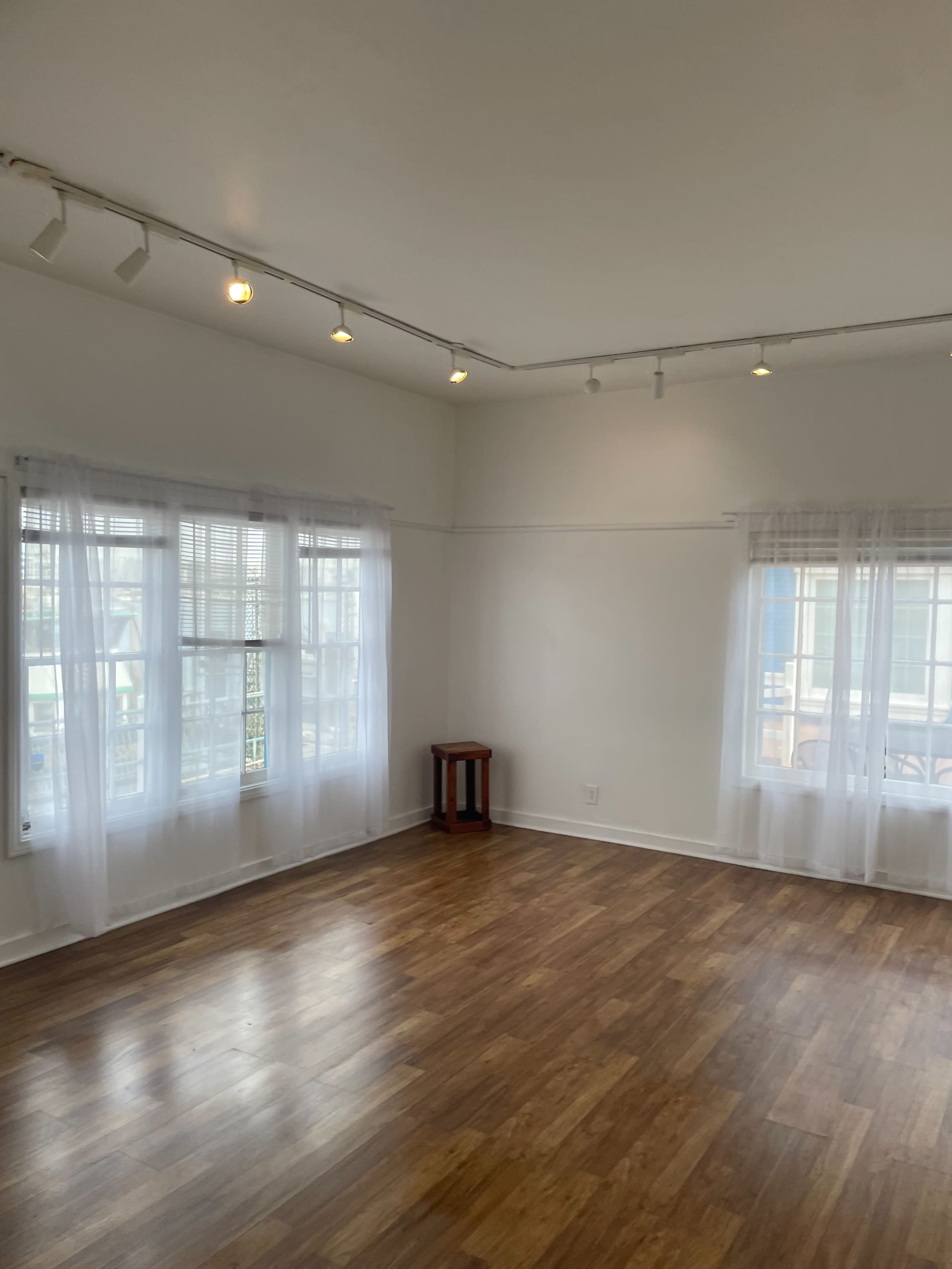 The image shows a bright, empty room with wooden floors, white walls, and large windows dressed with sheer curtains.