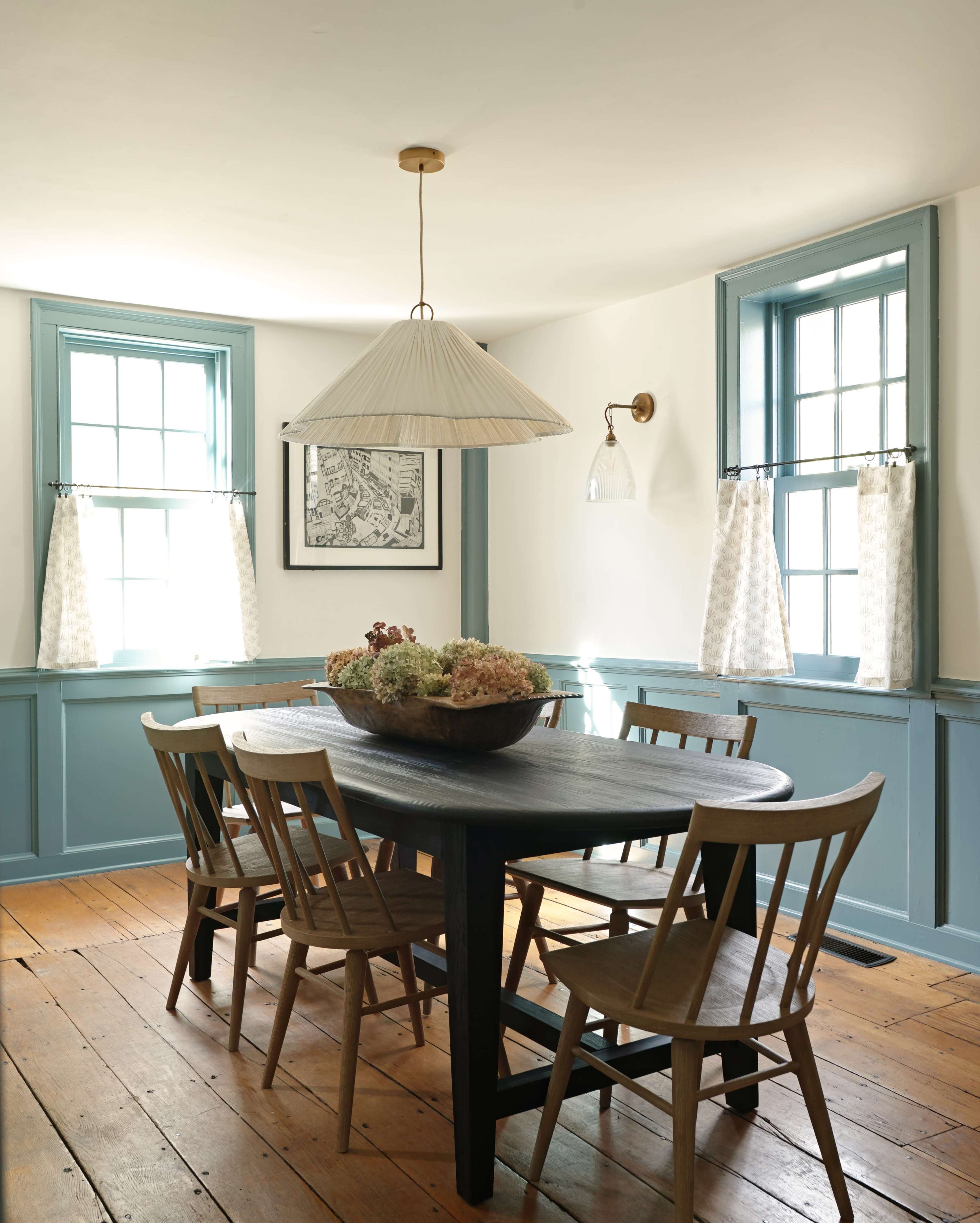 A dining room features a large round table surrounded by wooden chairs, illuminated by a pendant light, with windows dressed in light curtains and blue-painted walls.