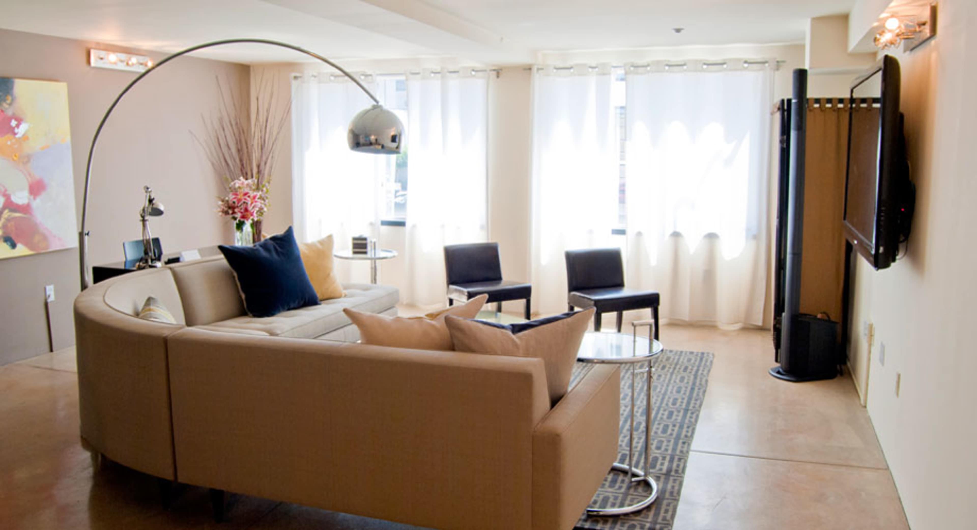 A modern living room with a curved sofa, a glass coffee table, and several chairs, illuminated by natural light through sheer curtains.