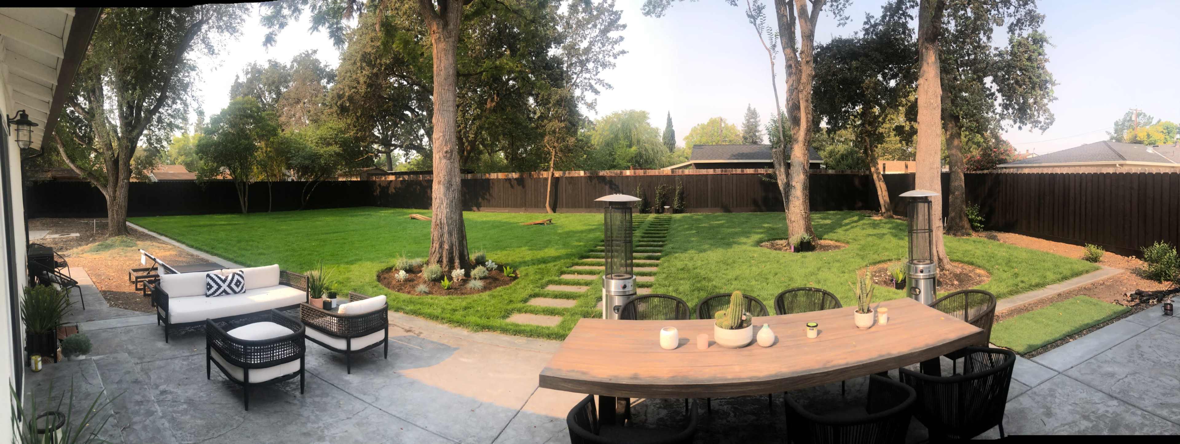 A panoramic view of a landscaped backyard featuring a large grassy area, a dining table set with decor, seating arrangements, and a pathway leading to a shaded area.