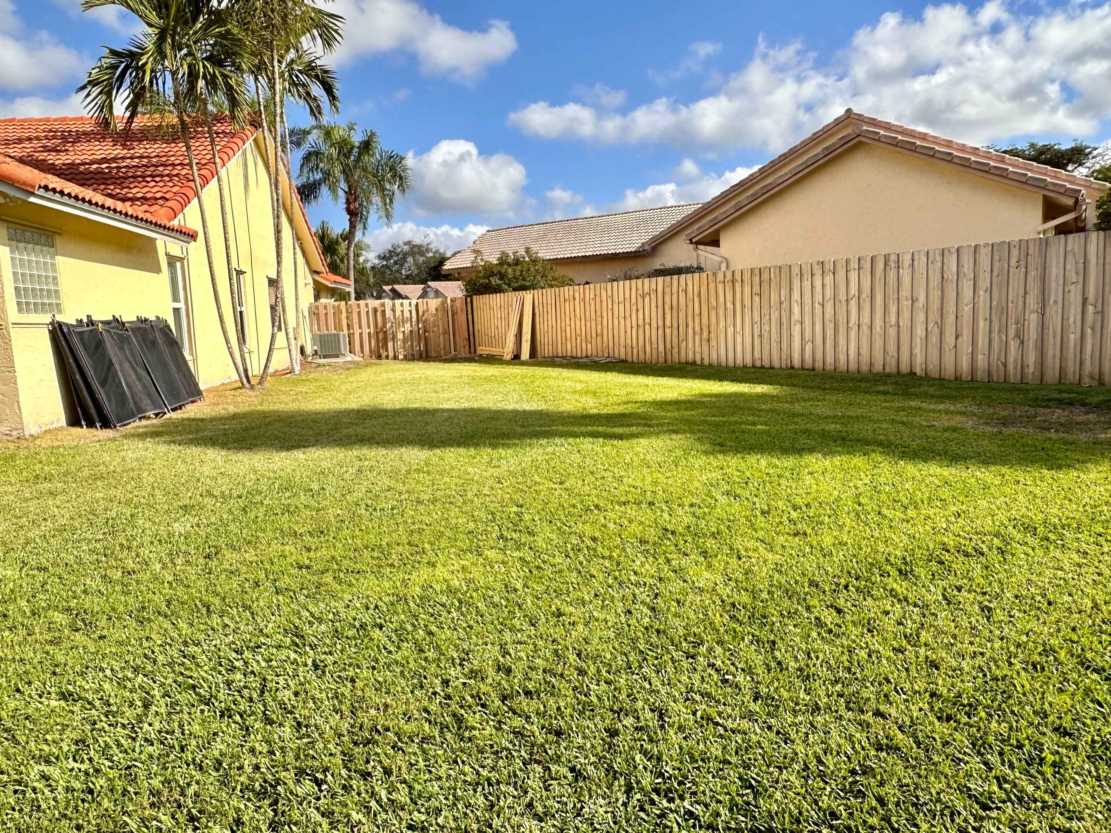 A grassy backyard enclosed by a wooden fence, with houses and palm trees in the background.