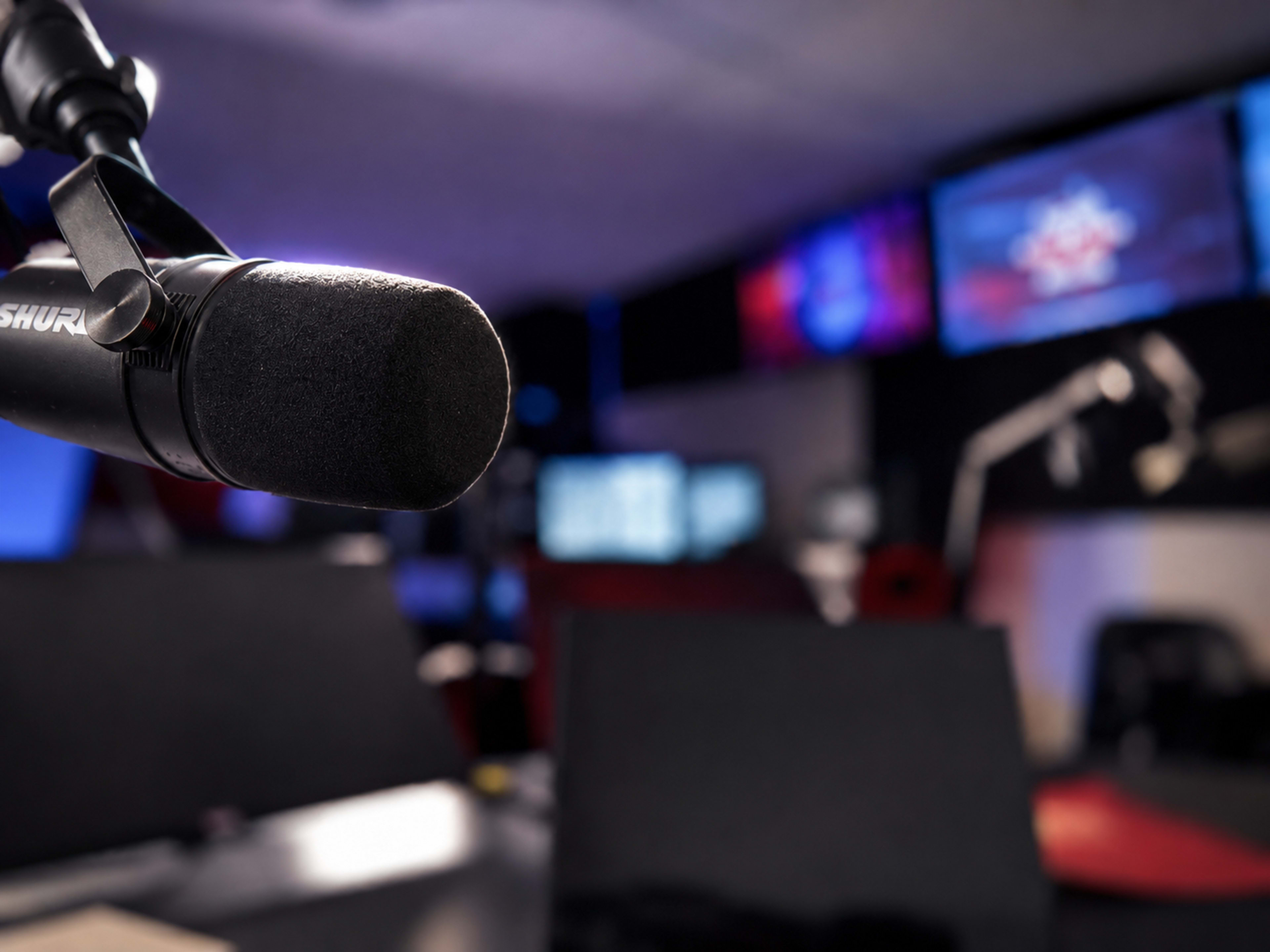 A close-up of a microphone is shown in a dimly lit studio with multiple screens in the background.