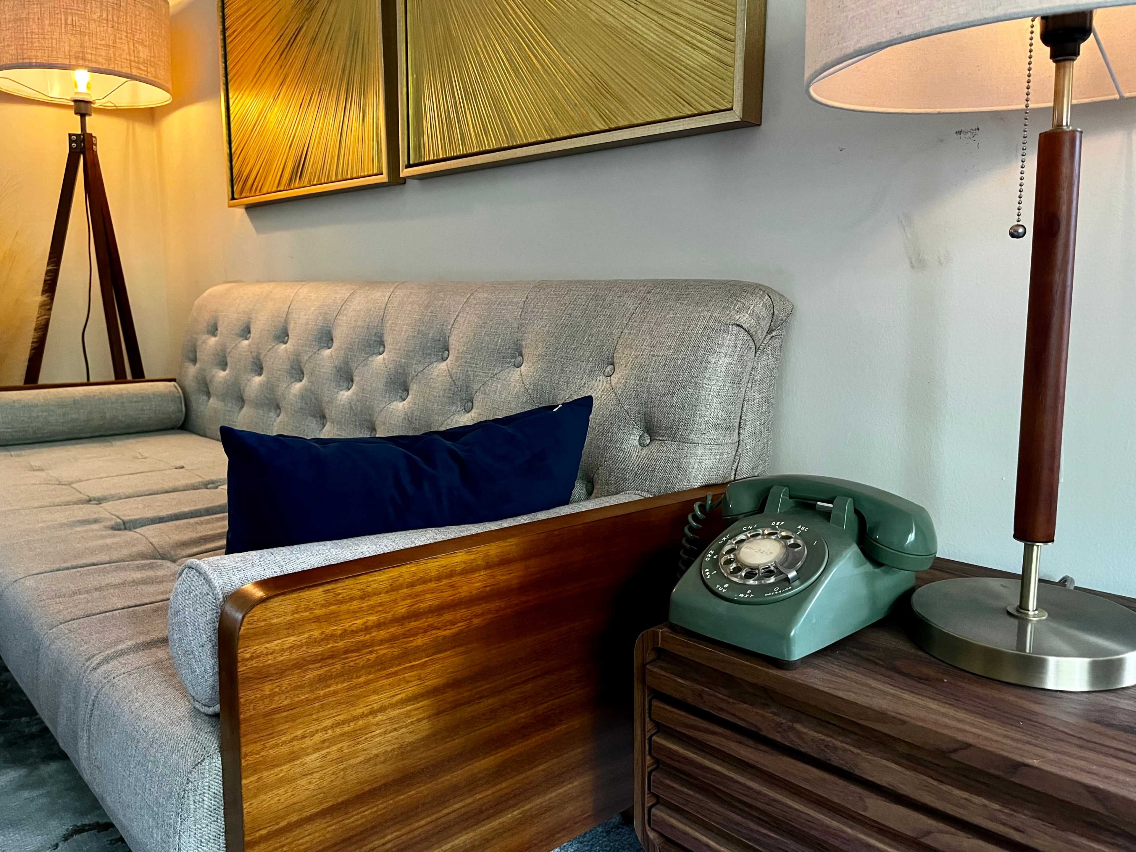 A modern living room scene with a light gray tufted sofa, a vintage green rotary phone on a wooden side table, and warm lighting from a standing lamp and a table lamp.
