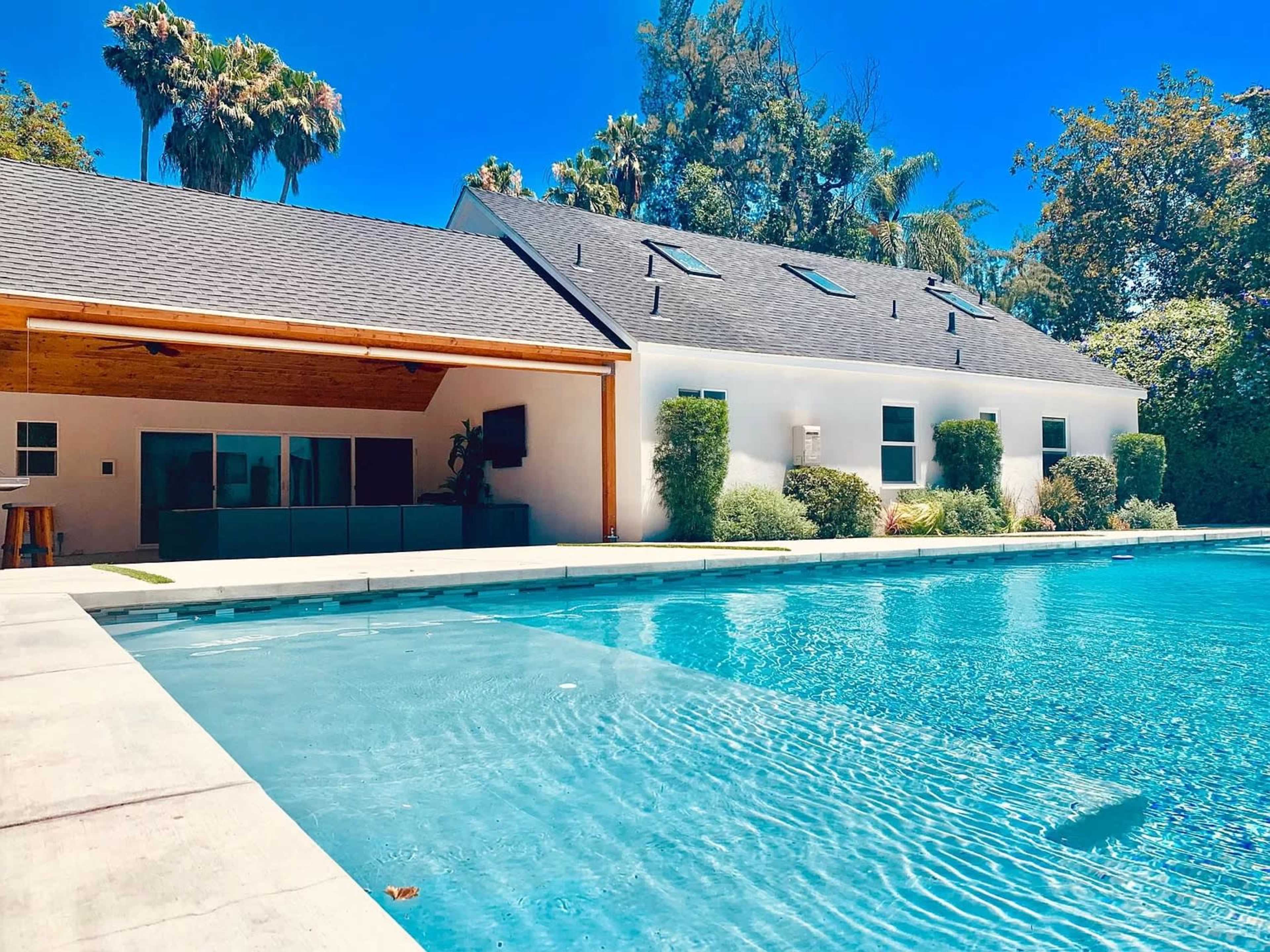 A swimming pool is situated next to a modern house with a sloped roof and landscaped greenery in the background.