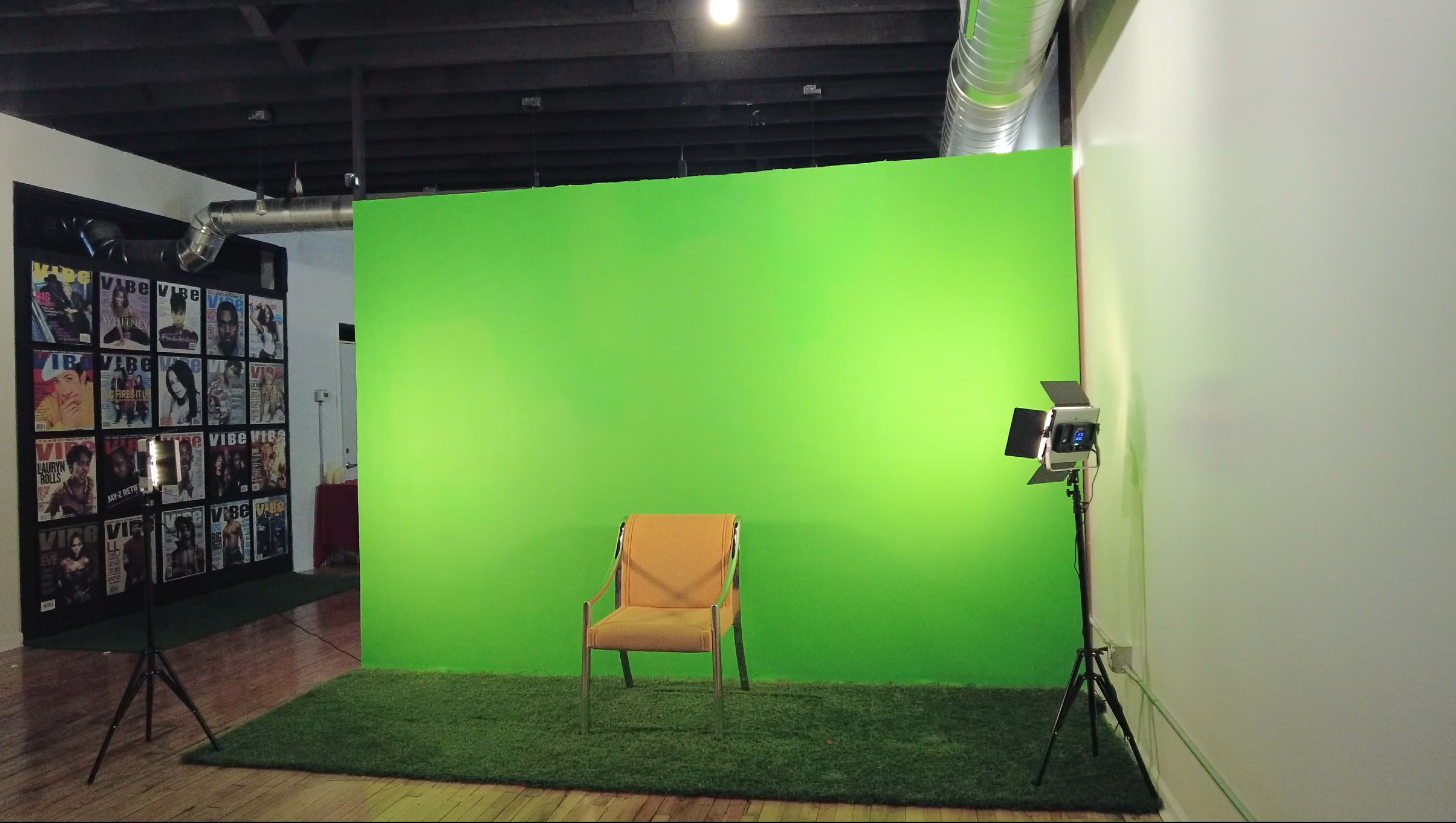 An empty orange chair positioned in front of a bright green backdrop, with a light setup on a stand beside it in a studio space.