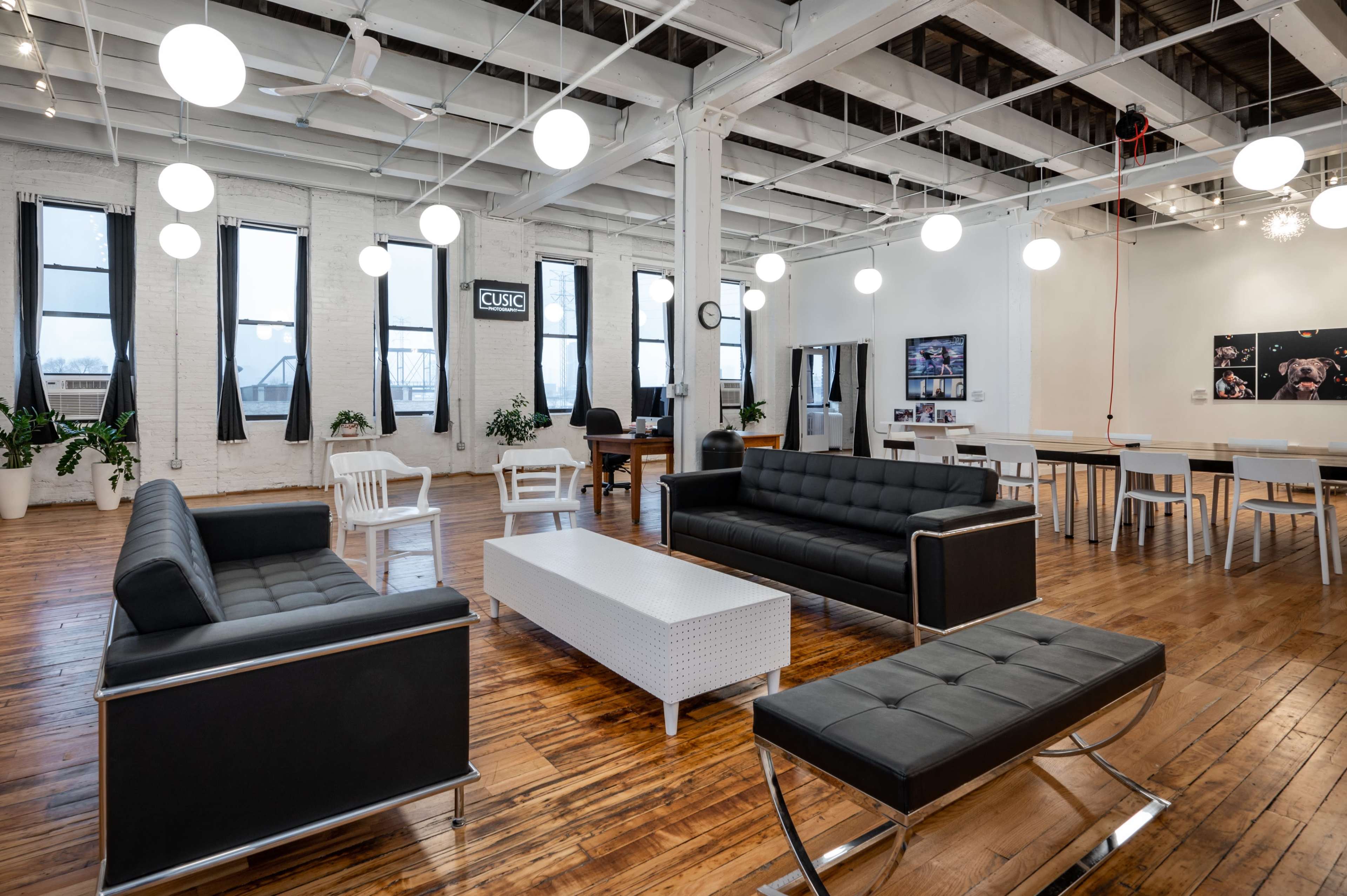 A modern, spacious lounge area with black sofas, a white coffee table, and a dining table surrounded by large windows and hanging lights.