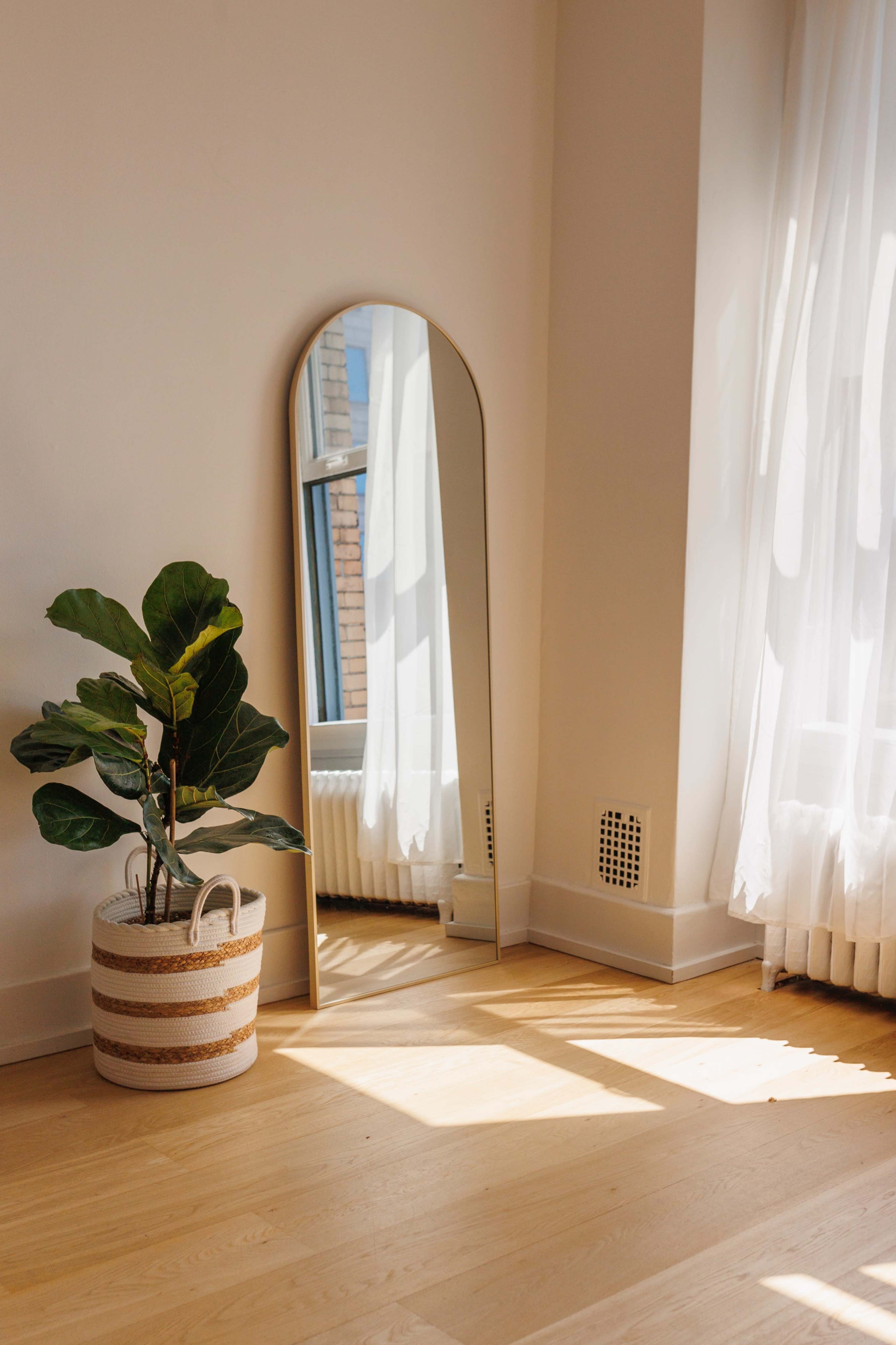A tall, arched mirror leans against a wall next to a potted plant in a decorative basket, with sunlight streaming through sheer curtains.