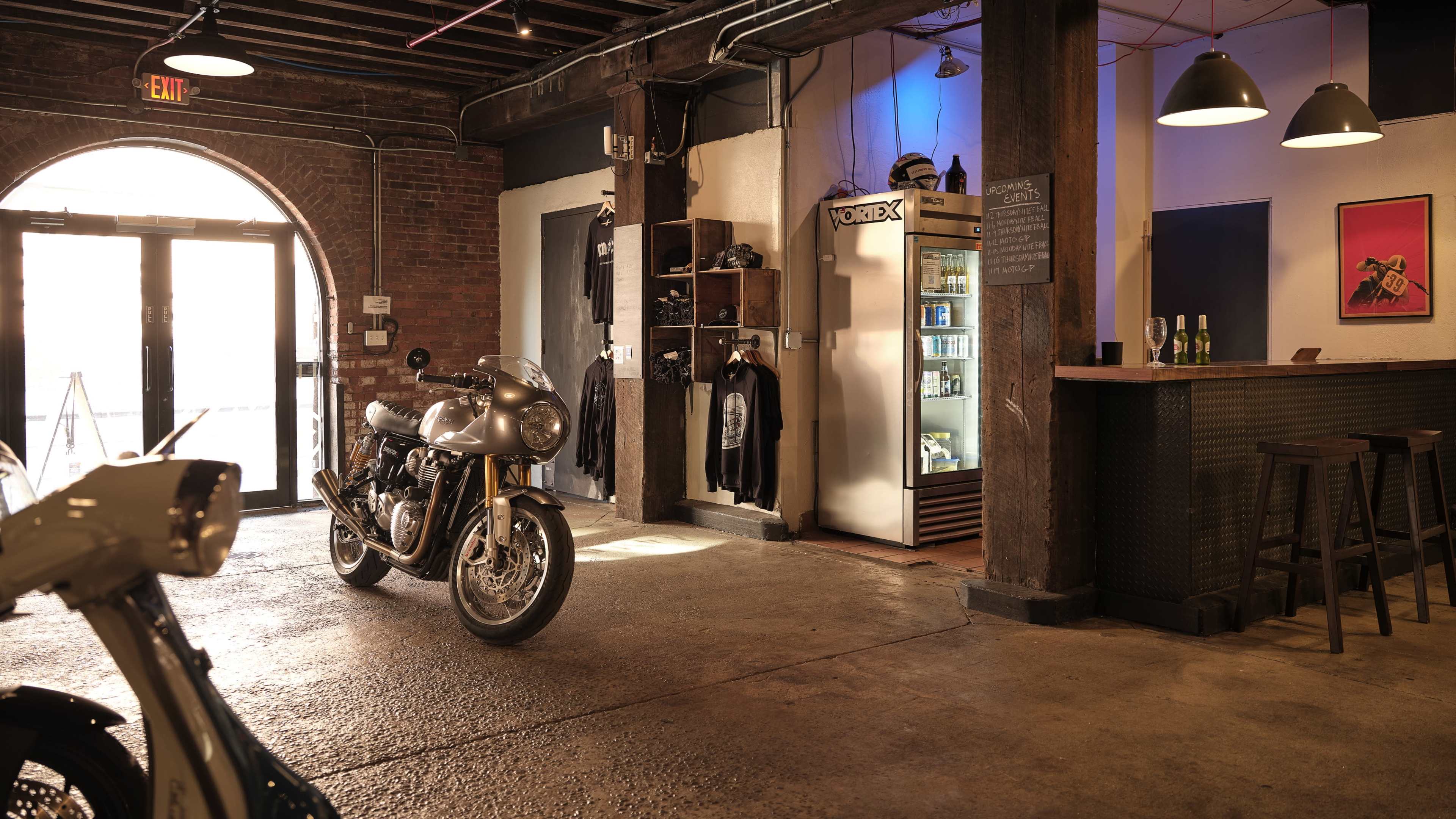 A spacious industrial-style interior featuring a classic motorcycle parked in front of a bar area, with clothing displays and a vending fridge along the walls.