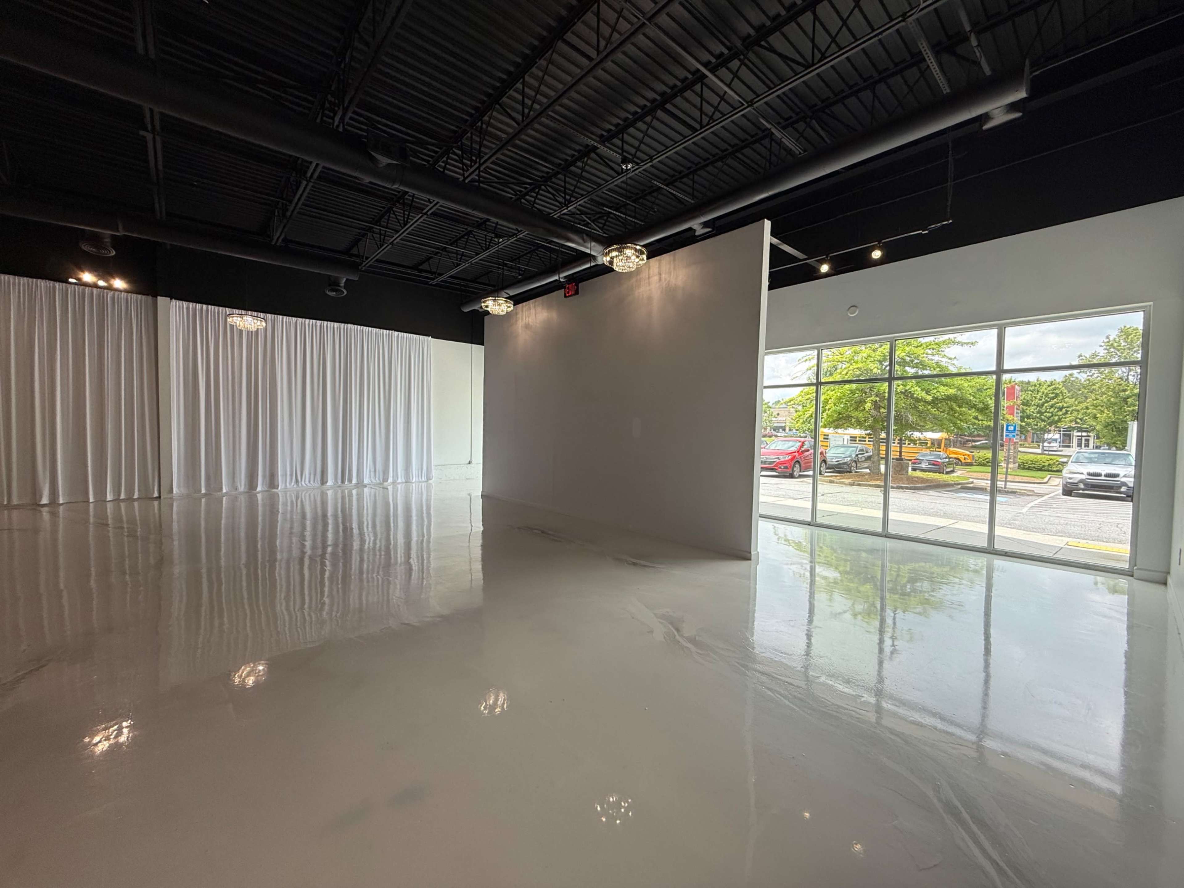 The image shows a spacious, empty interior with polished white floors, draped white curtains on one side, and large glass windows overlooking an outdoor area.