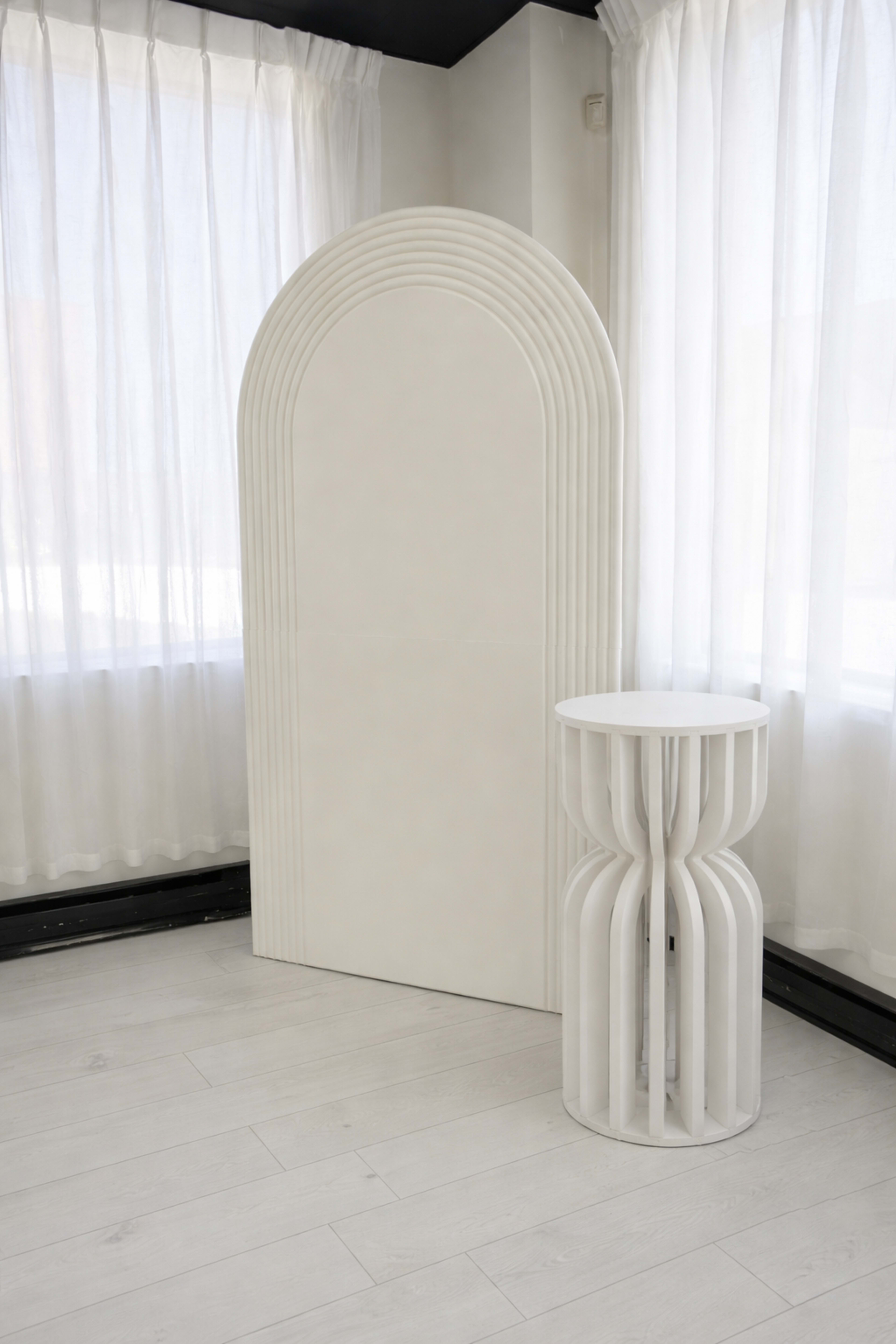The image features a minimalist interior with a white, arch-shaped backdrop and a cylindrical side table, both illuminated by natural light from large windows.