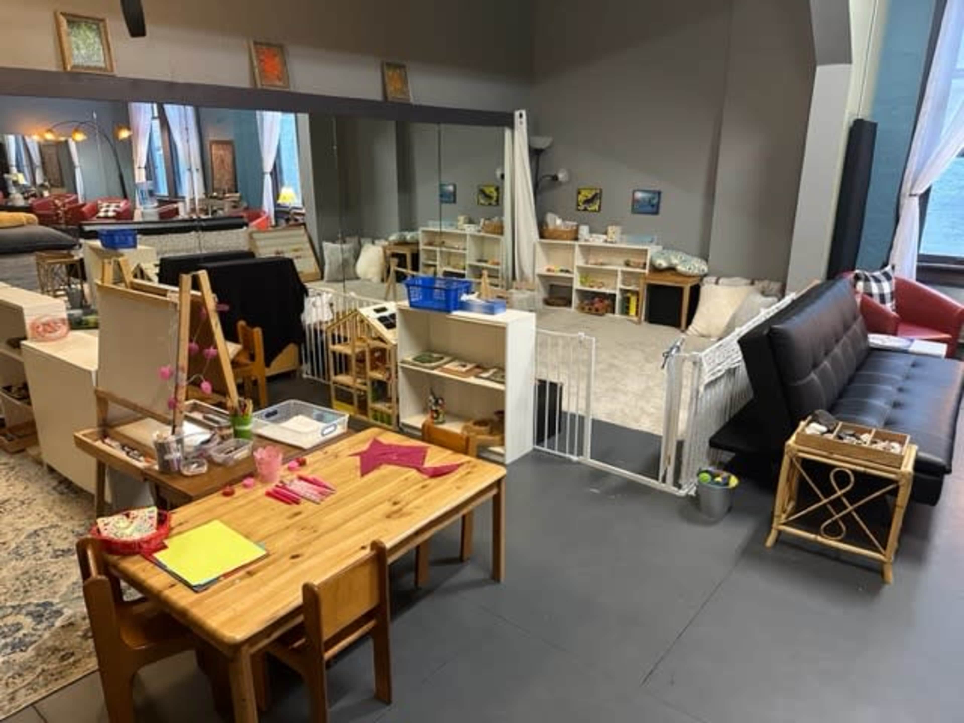The image shows a spacious room with a children's play area featuring a wooden table and chairs, a black sofa, and shelves filled with toys.