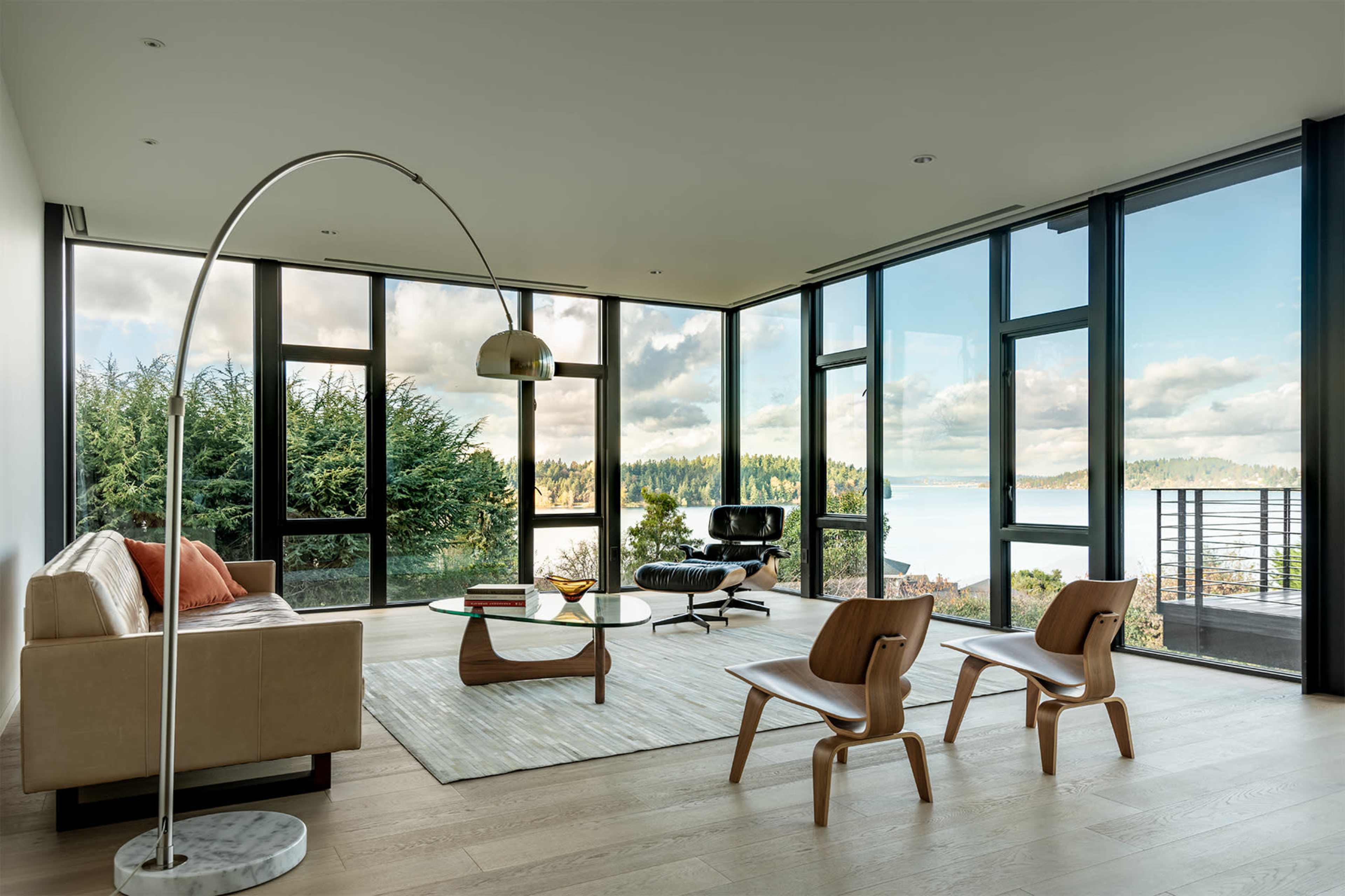 A modern living room features large floor-to-ceiling windows with scenic views, minimalist furniture, and a neutral color palette.