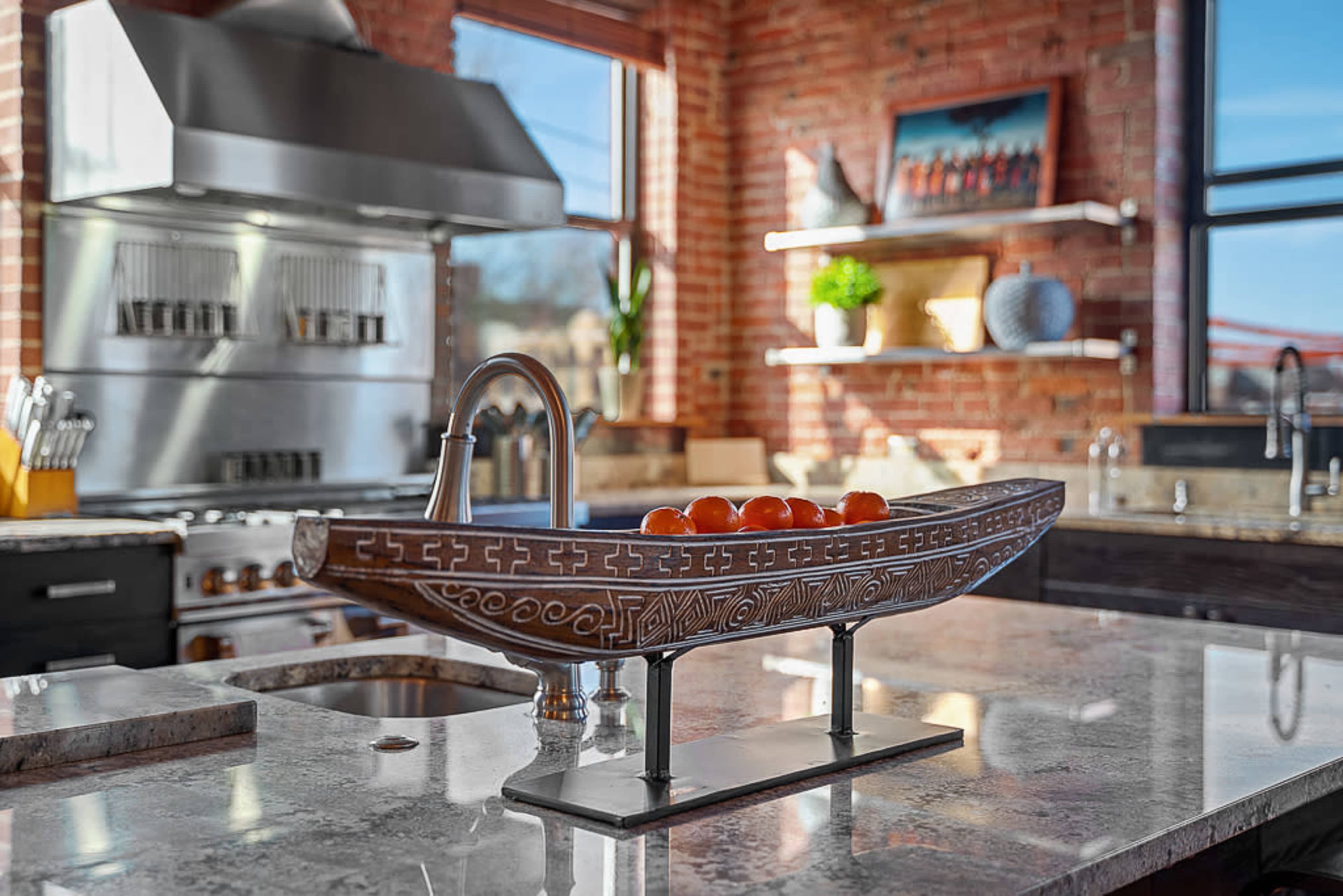 A modern kitchen features a decorative wooden bowl filled with fruit on a granite countertop, alongside stainless steel appliances and exposed brick walls.
