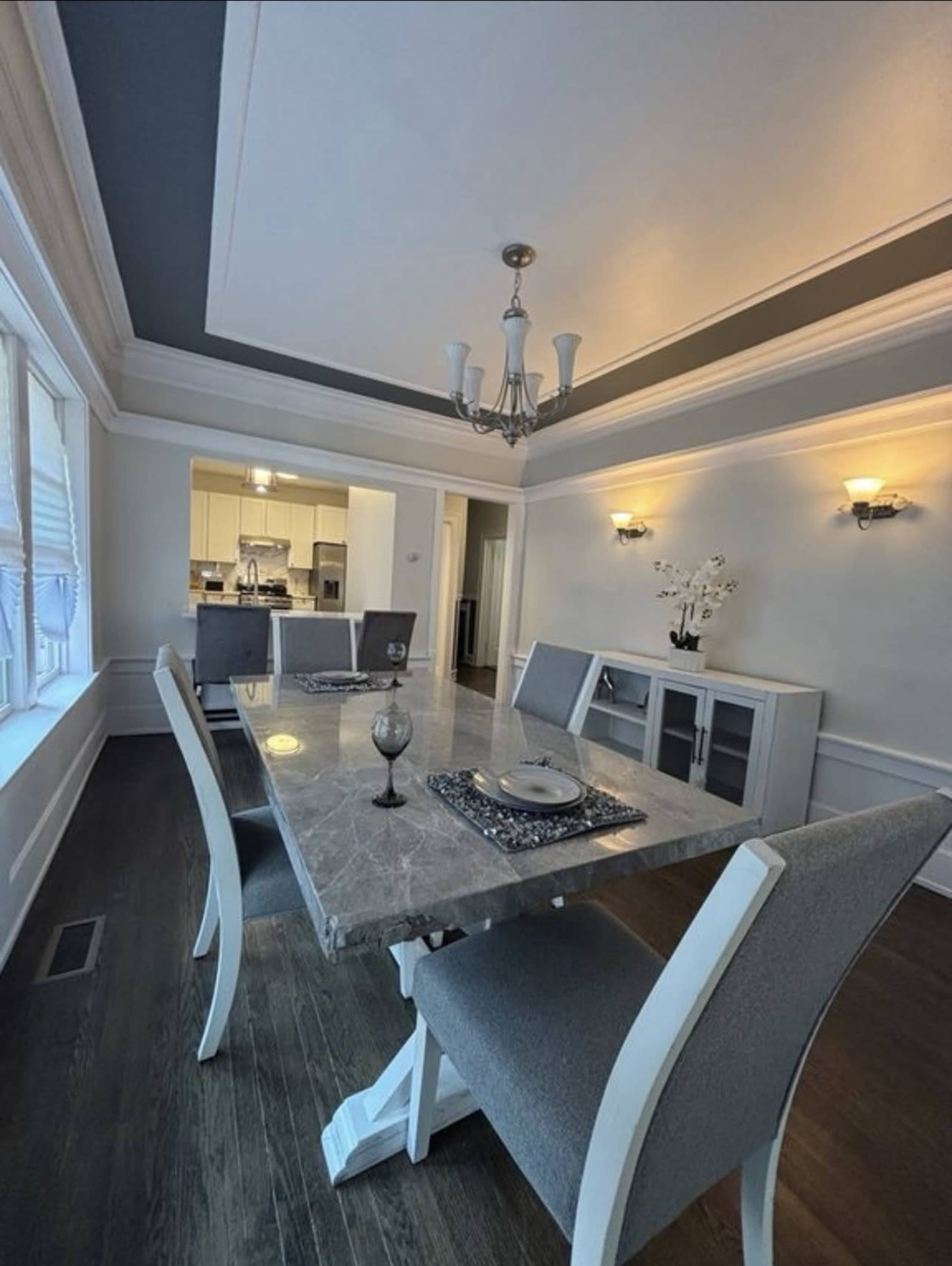A dining room features a large marble table surrounded by gray upholstered chairs and a chandelier above.